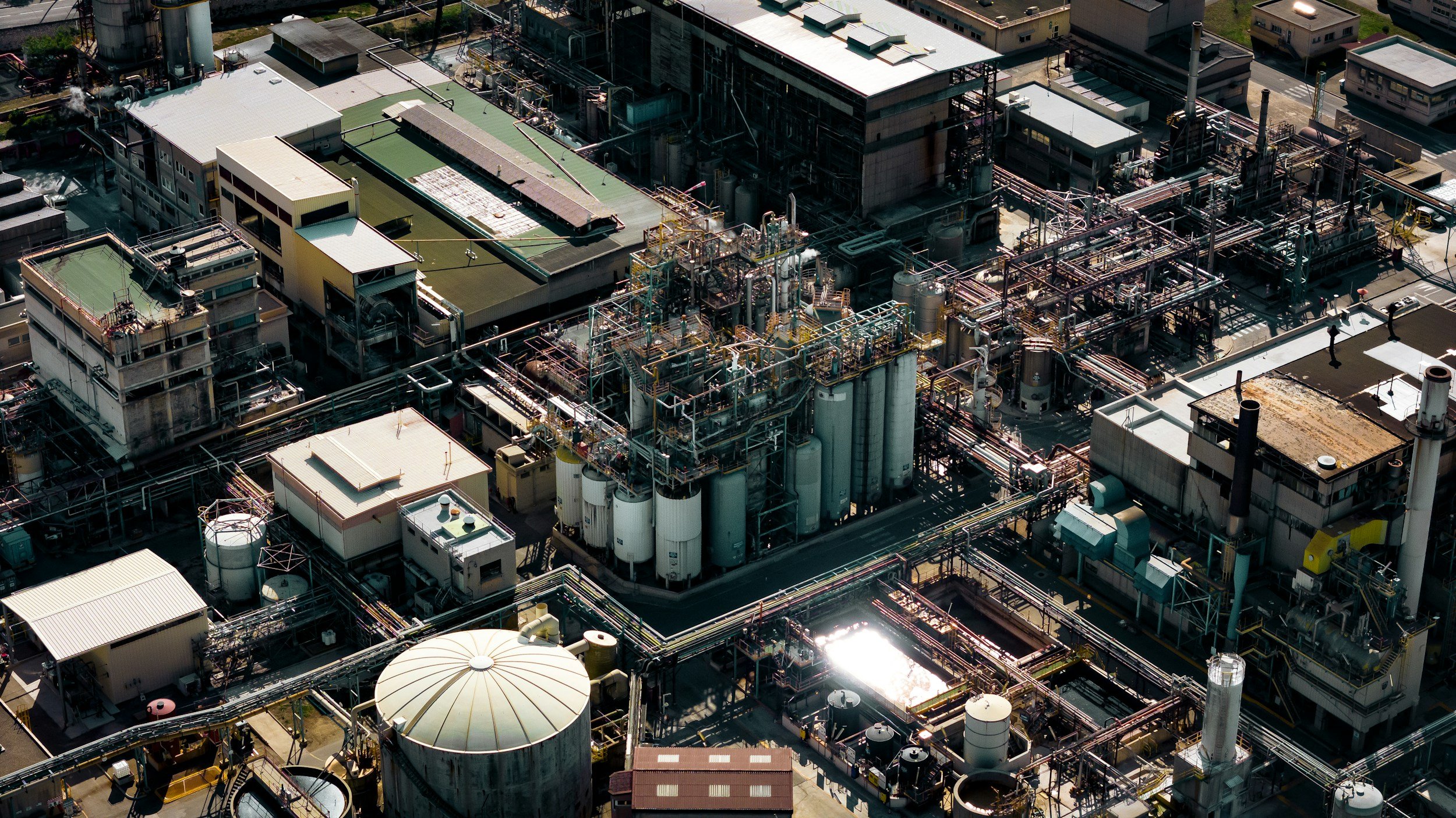 Aerial view of an industrial factory with interconnected pipes, storage tanks, and buildings, surrounded by residential houses.