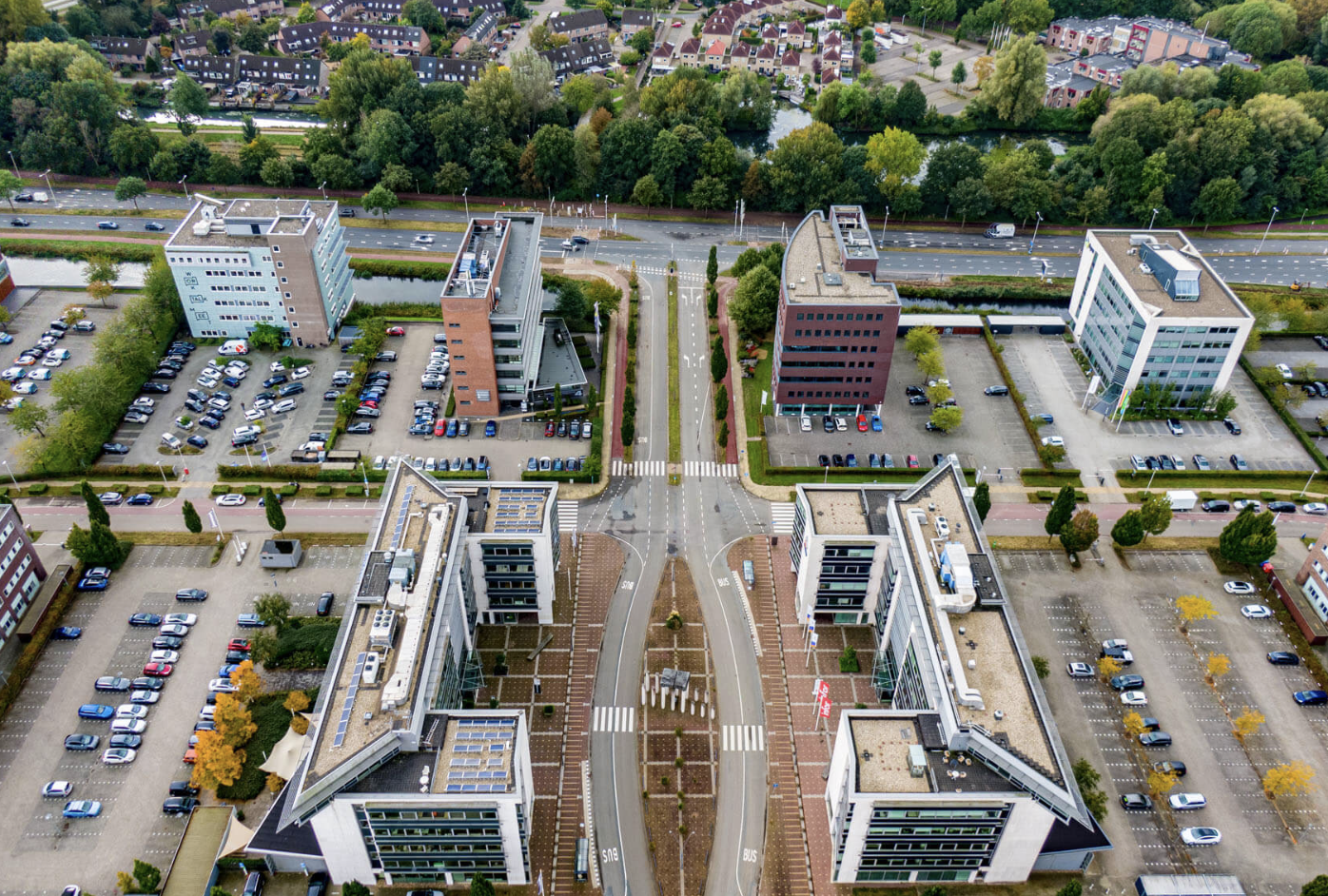 An aerial view of a modern cityscape with several buildings, parking lots, and a street with cars and trees, adjacent to a green park with water features.