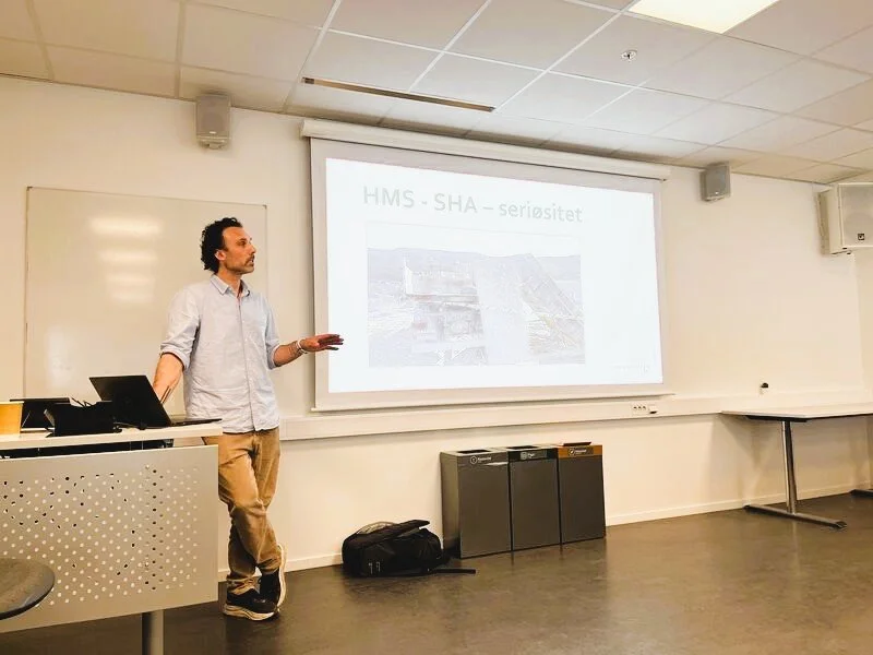 A man standing in a classroom giving a presentation with a slide projected on a whiteboard behind him. The slide has text that reads 'HMS - SHA - seriositet' and shows some images.
