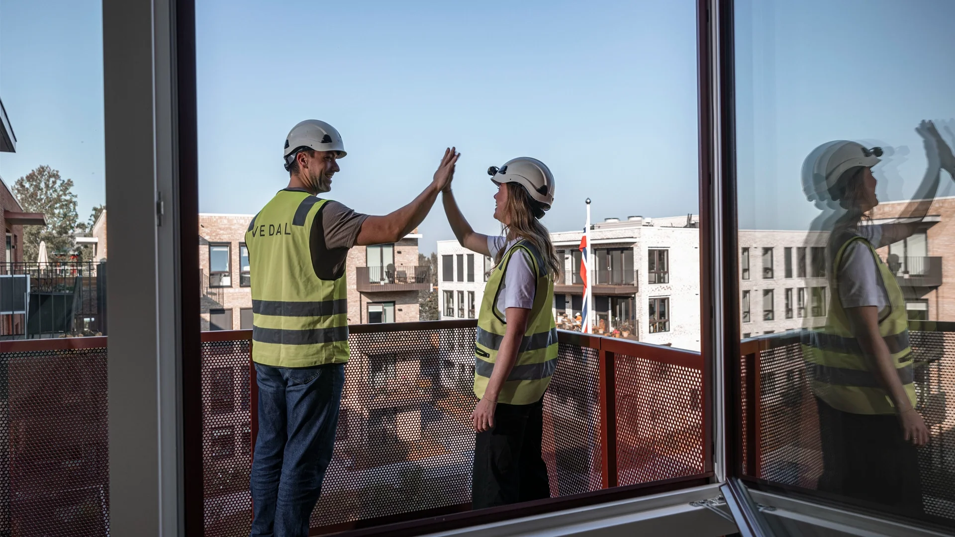 A construction worker man and woman high-fiving on a balcony with a background of modern apartment buildings and a clear blue sky.