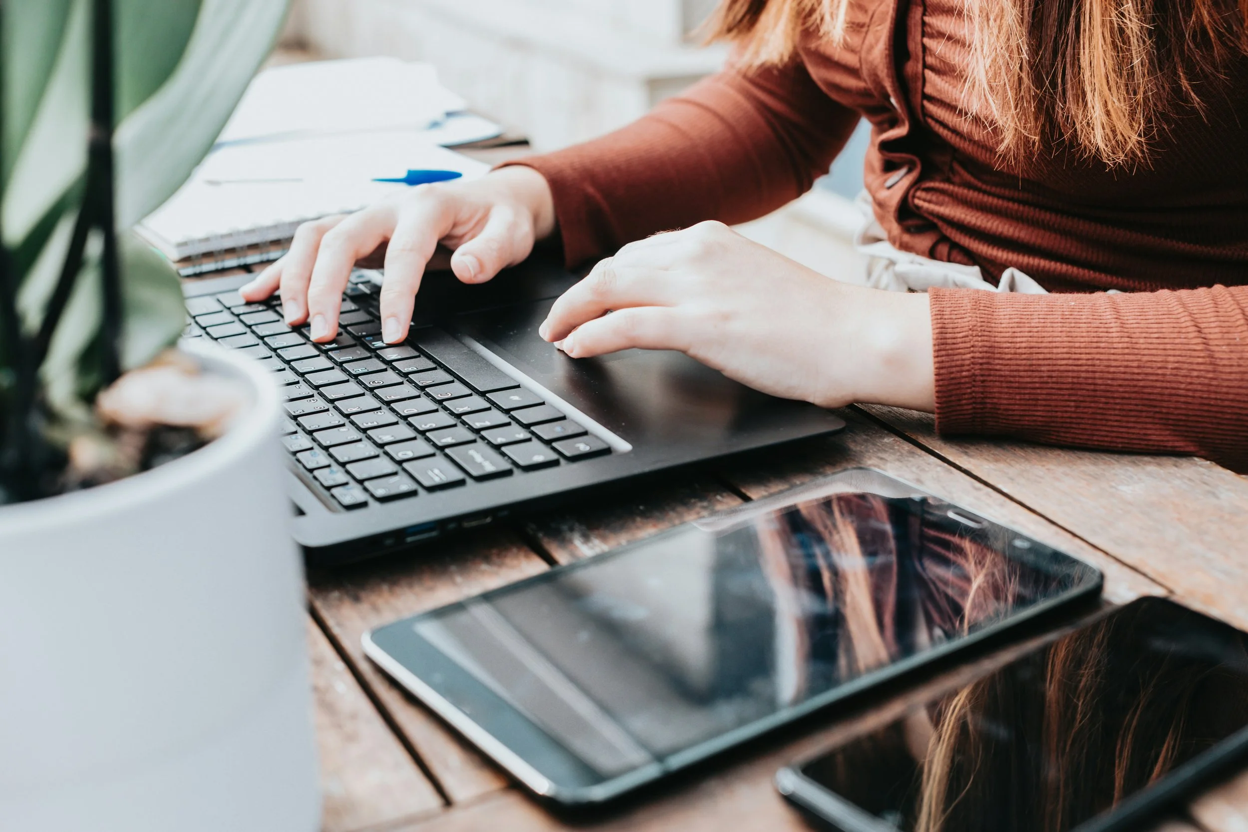 Person typing on a laptop with a notebook, pen, smartphone, and plant on a wooden table.