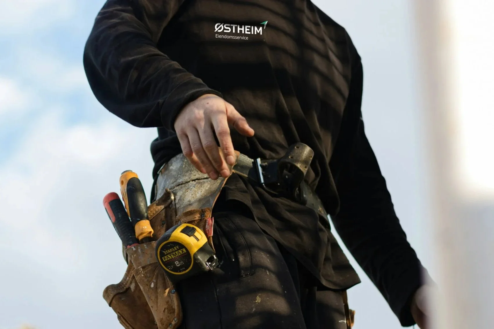 A person dressed in dark work clothes with a logo on the chest, wearing a tool belt with various tools, adjusting a piece of material, likely siding, on a construction site or roof against the sky.
