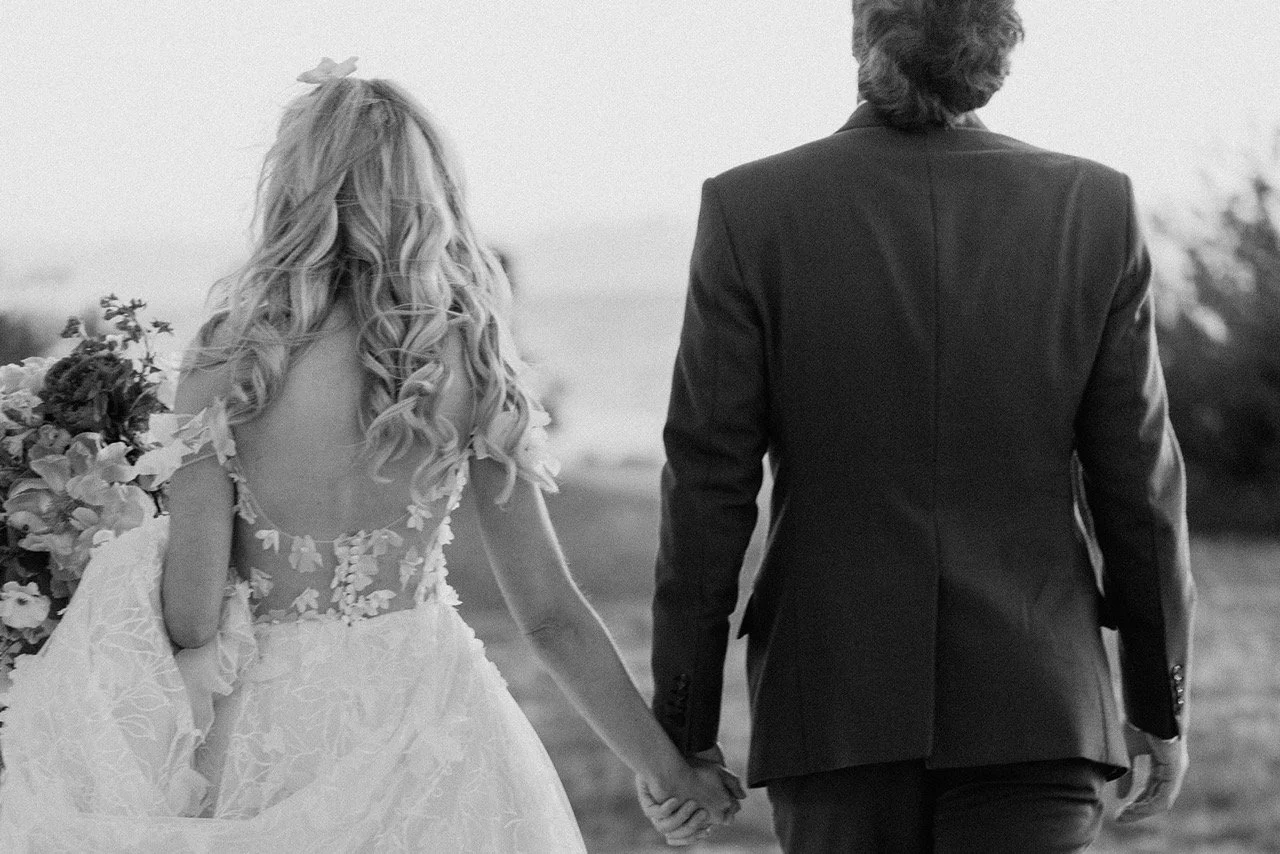 Pareja en traje de boda, la mujer con vestido y el hombre con traje, caminando de la mano en un exterior.