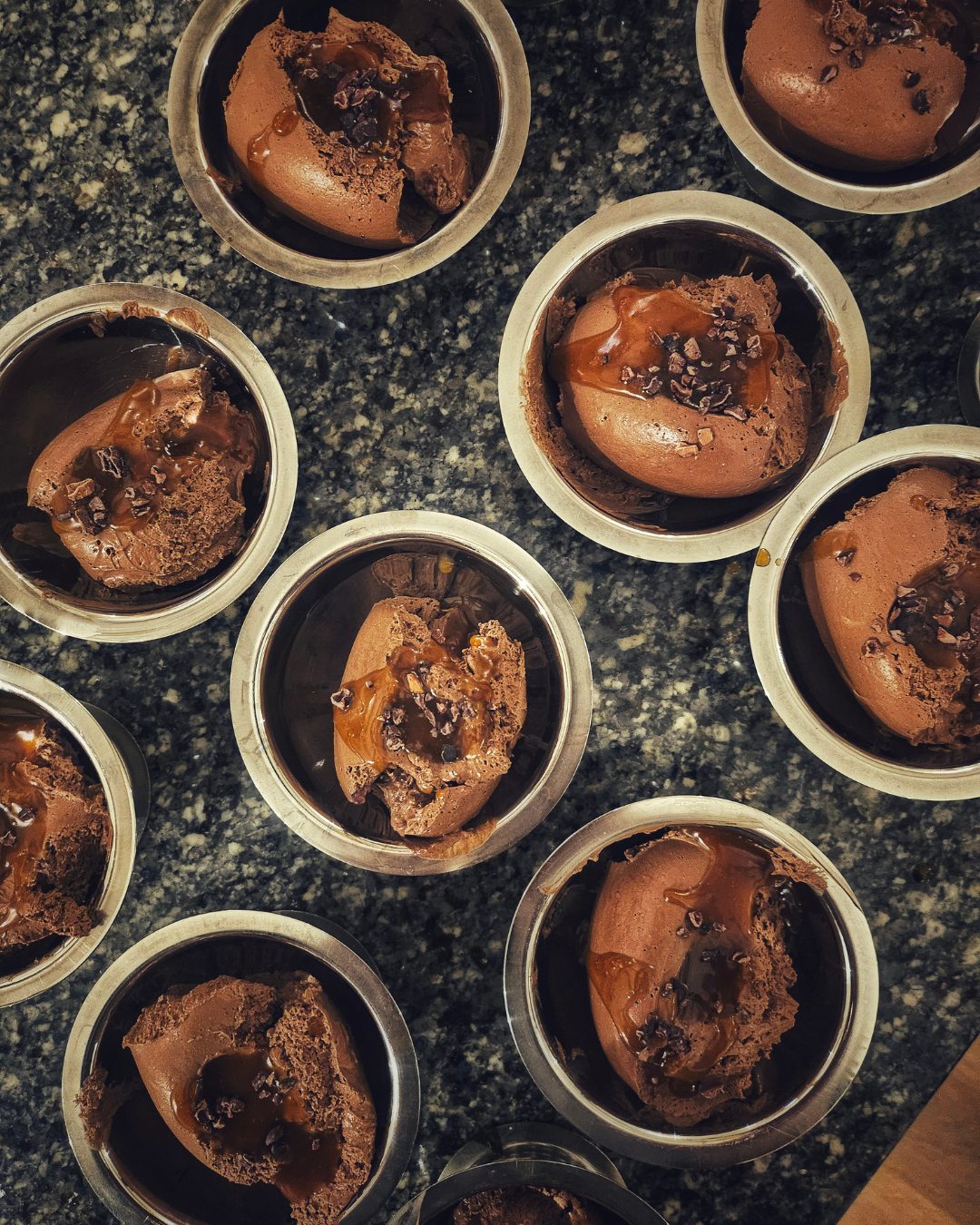 Multiple cups of chocolate mousse with chocolate shavings and caramel, served on a dark granite countertop.