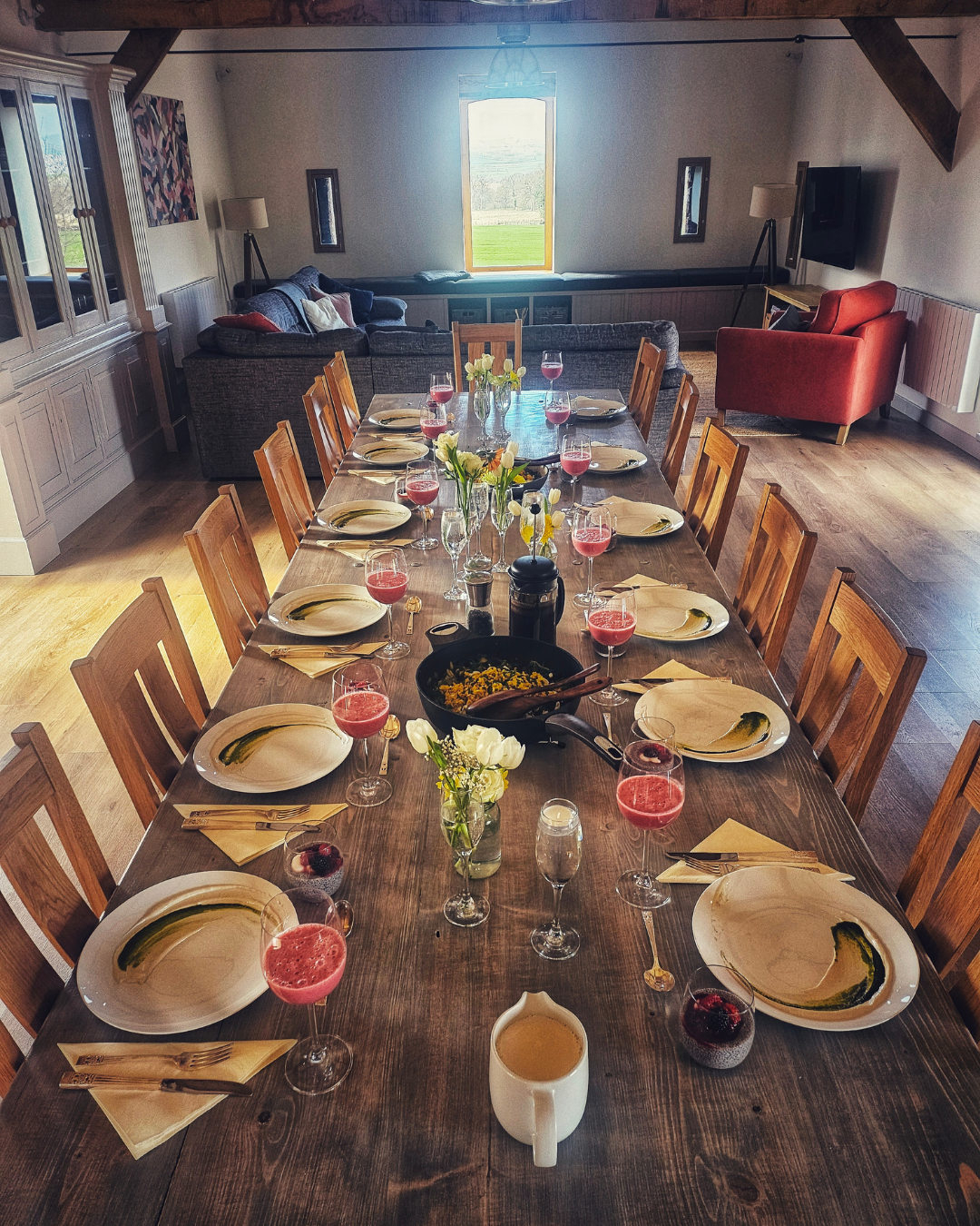 A large wooden dining table set for a meal with twelve place settings, including plates, glasses with pink and clear drinks, and yellow napkins with utensils. There are small flower arrangements and a large skillet with food in the center. In the bac