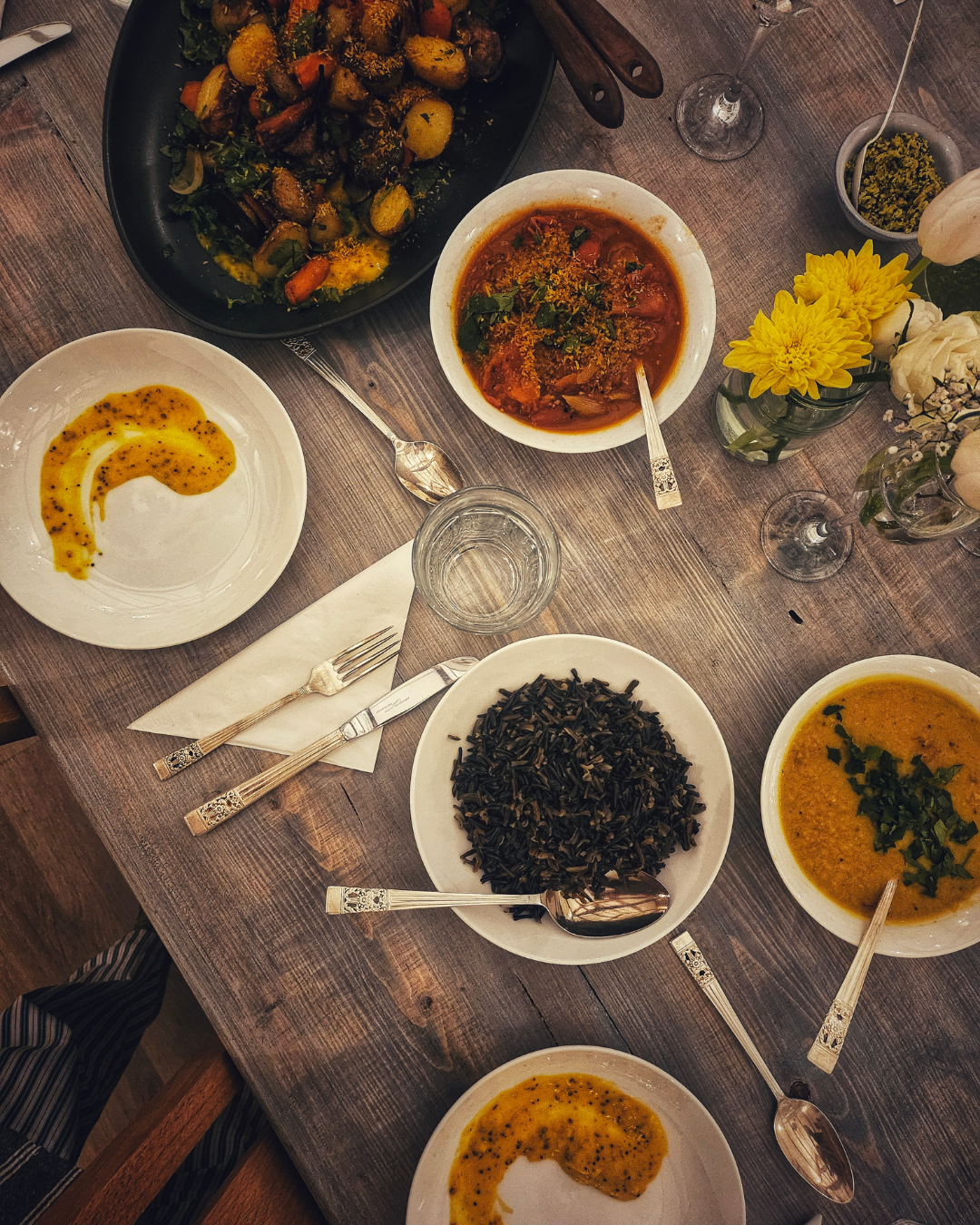 A wooden table set with various dishes including rice, vegetable stir-fry, vegetable soup, and a tomato-based stew, along with wine glasses, cutlery, and a vase of mixed flowers.