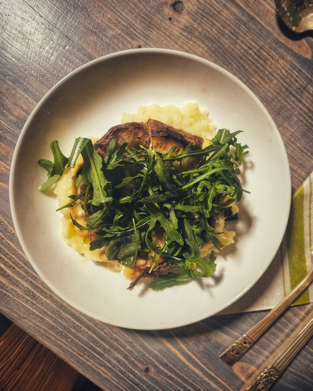A white plate with mashed potatoes, grilled meat, and a green arugula salad on top, placed on a wooden table.