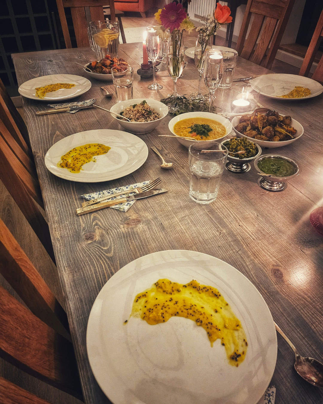 A dinner table set with various dishes including bowls of lentil soup, rice, roasted vegetables, and green sauces, along with glasses of water, champagne, and flowers for decoration.