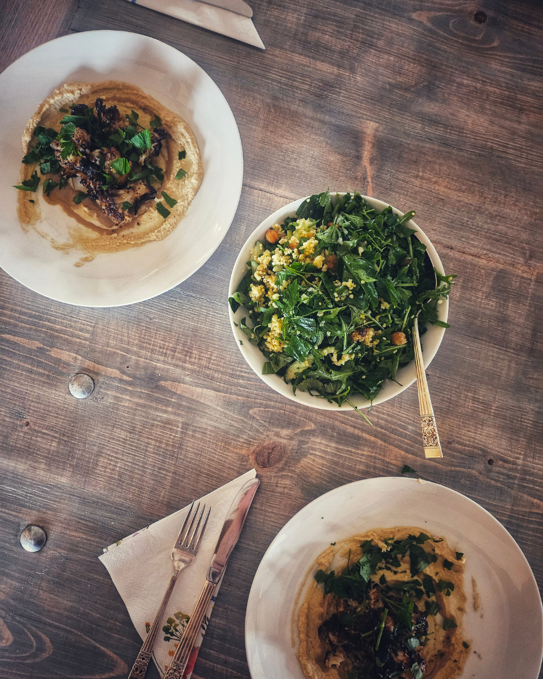 A wooden table with two plates of hummus topped with caramelized onions and herbs, and a bowl of fresh green salad with herbs, tomatoes, and nuts.