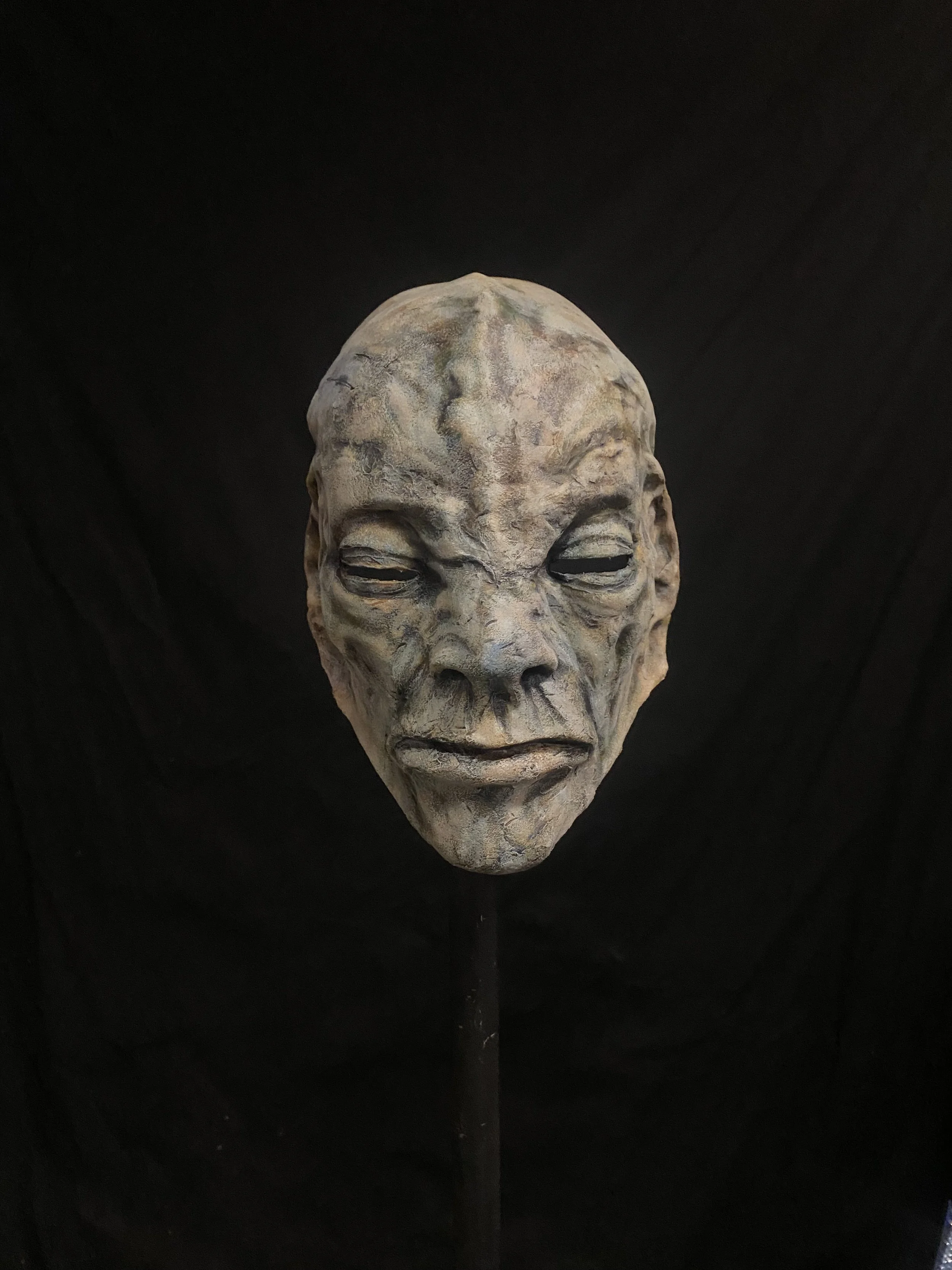 A sculpture of a human face with a weathered and textured surface, placed against a black background.