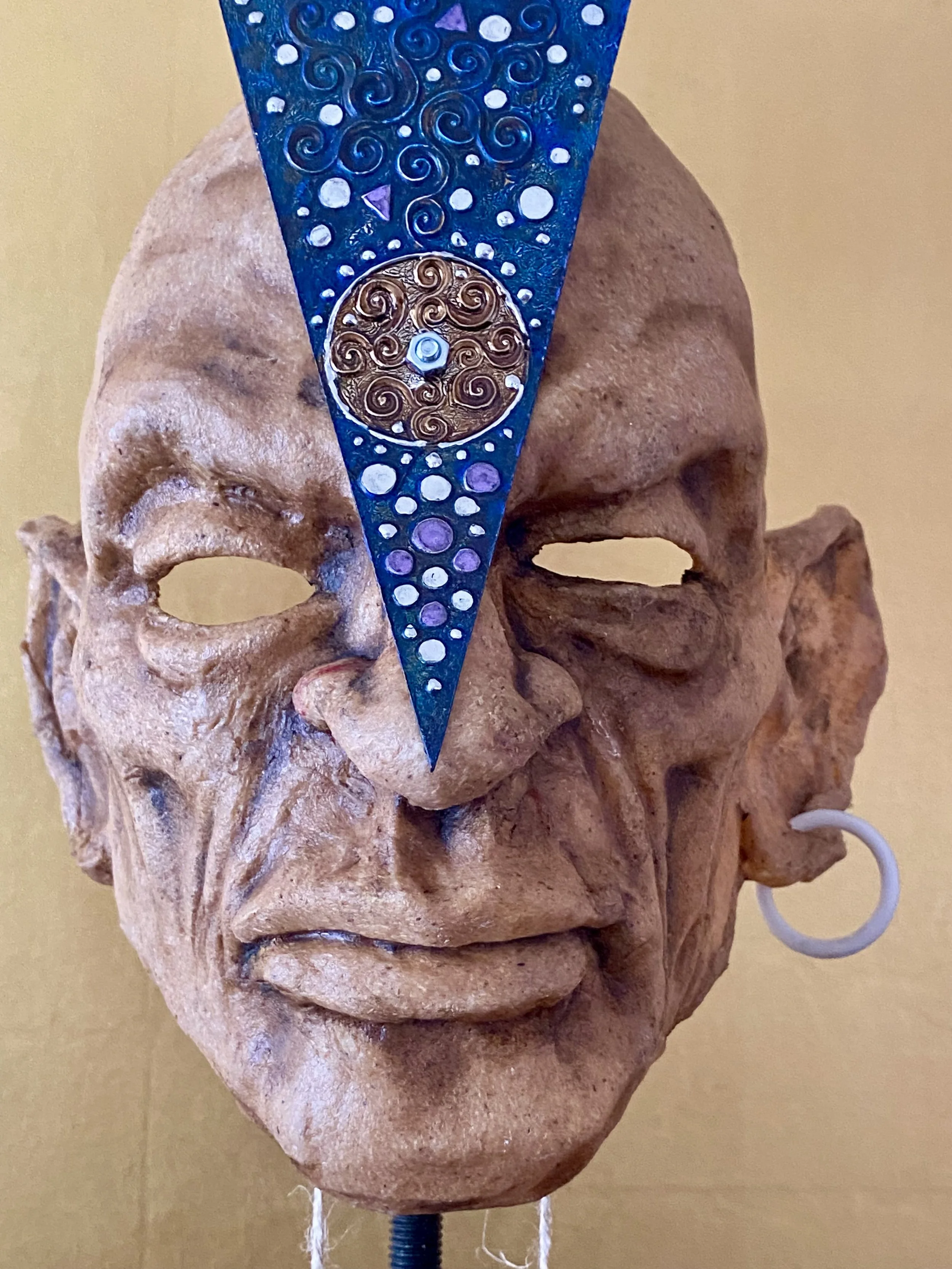 A carved clay or ceramic face mask with a metallic blue and gold decorative element covering the eye area, featuring swirling patterns and a circular design.