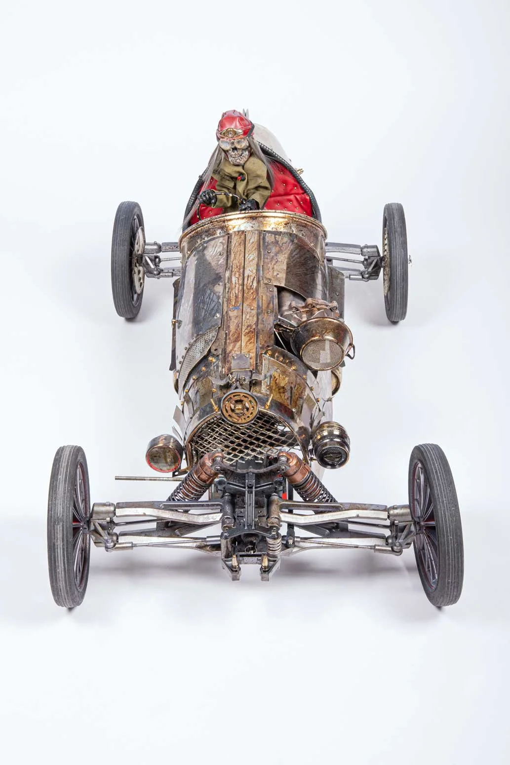 A vintage race car with a skeleton driver wearing a red helmet and goggles inside. The car has a rusted, weathered metal body with exposed mechanical parts and four wheels.