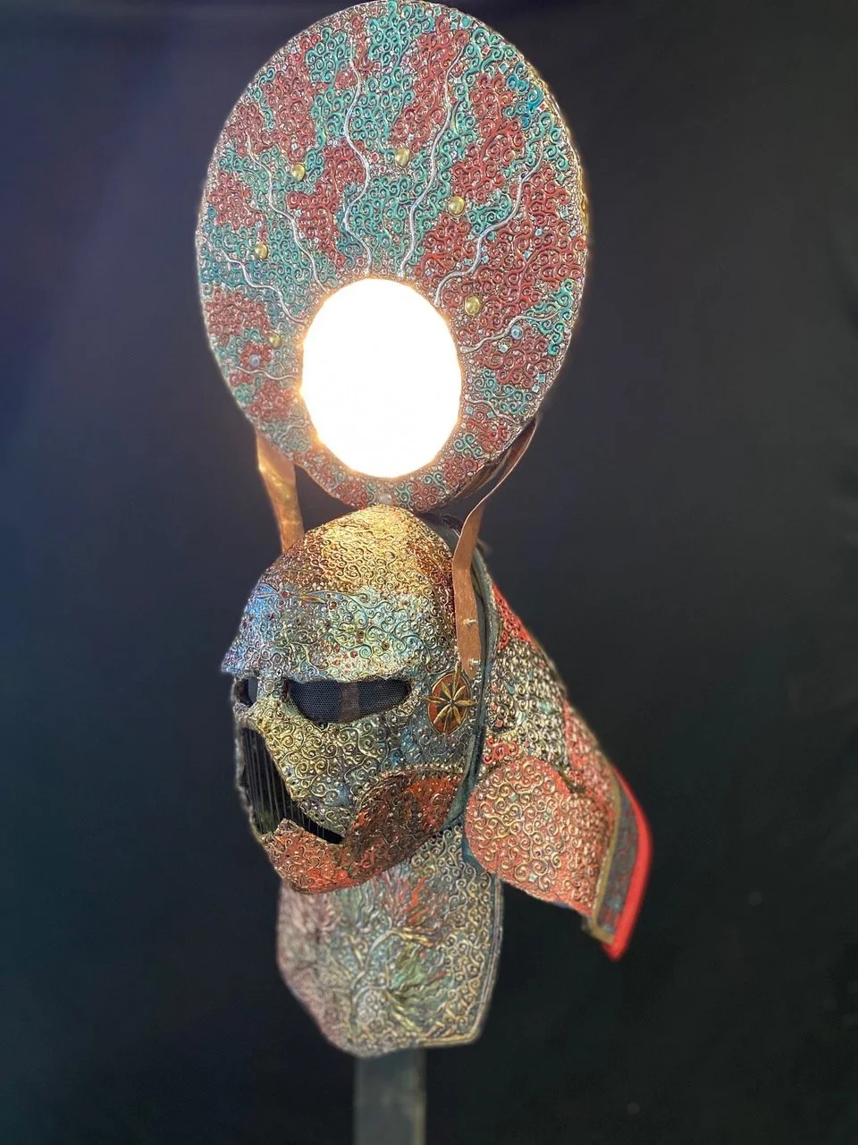 A decorative sorcerer  mask with an illuminated halo above a skull, featuring intricate, colorful patterns and a textured surface.