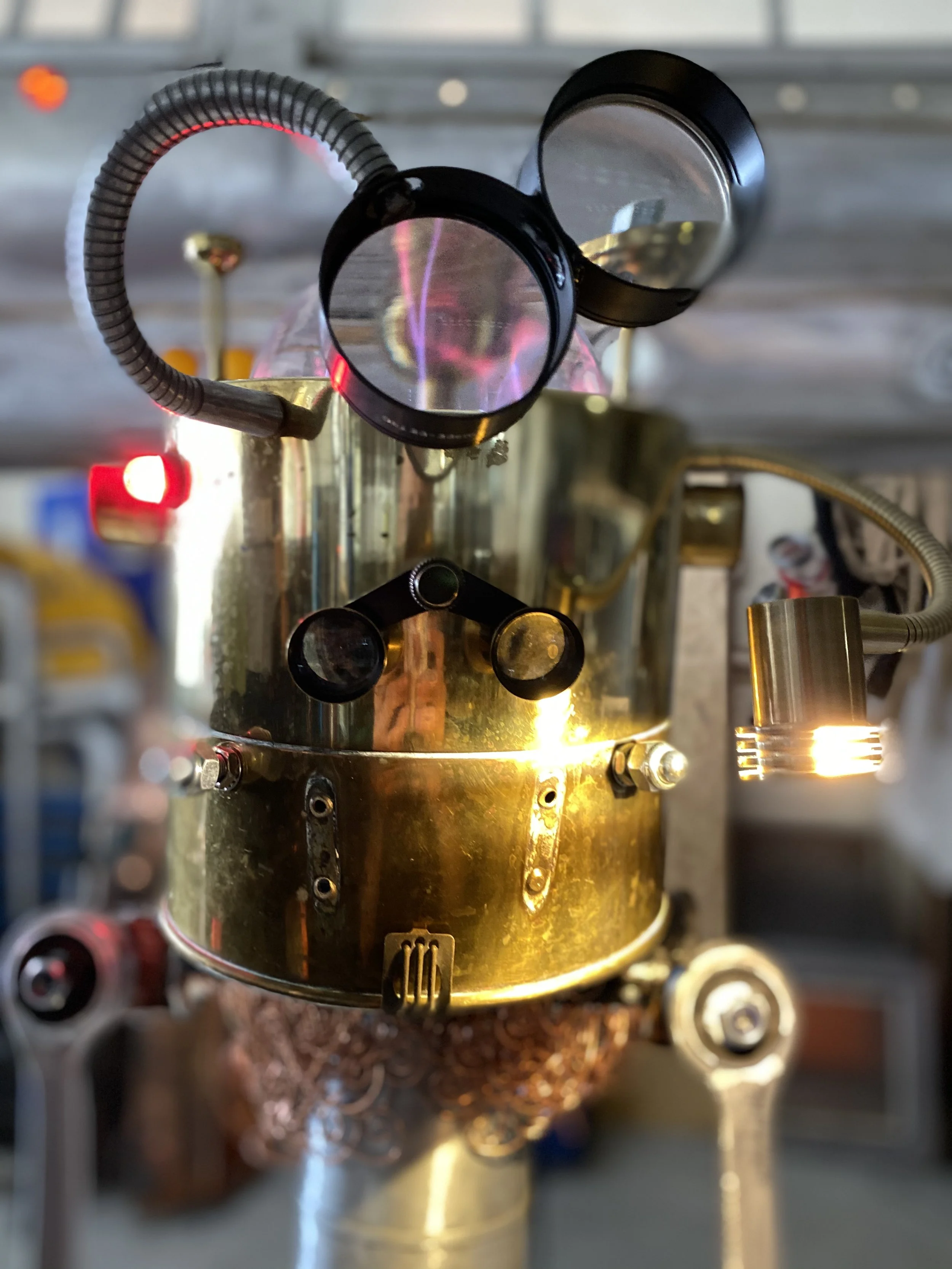 Close-up of a vintage robotic machine with metallic and brass components, circular lenses, a flexible hose, and a copper coil underneath, set in a workshop environment.