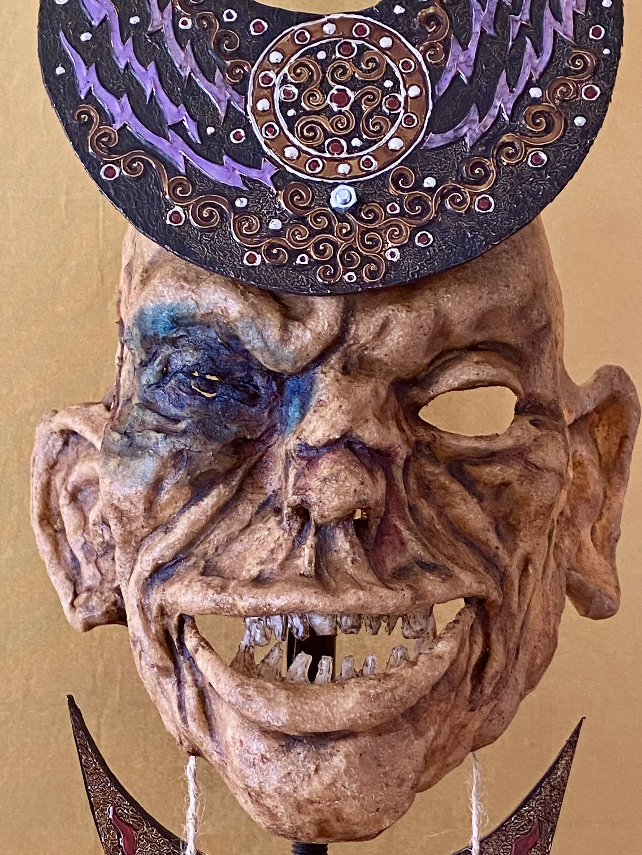 A grotesque and detailed mask resembling a smiling, aged man with prominent wrinkles, missing teeth, and a large nose. The mask is topped with a decorative, circular, black and purple object with intricate swirling patterns.