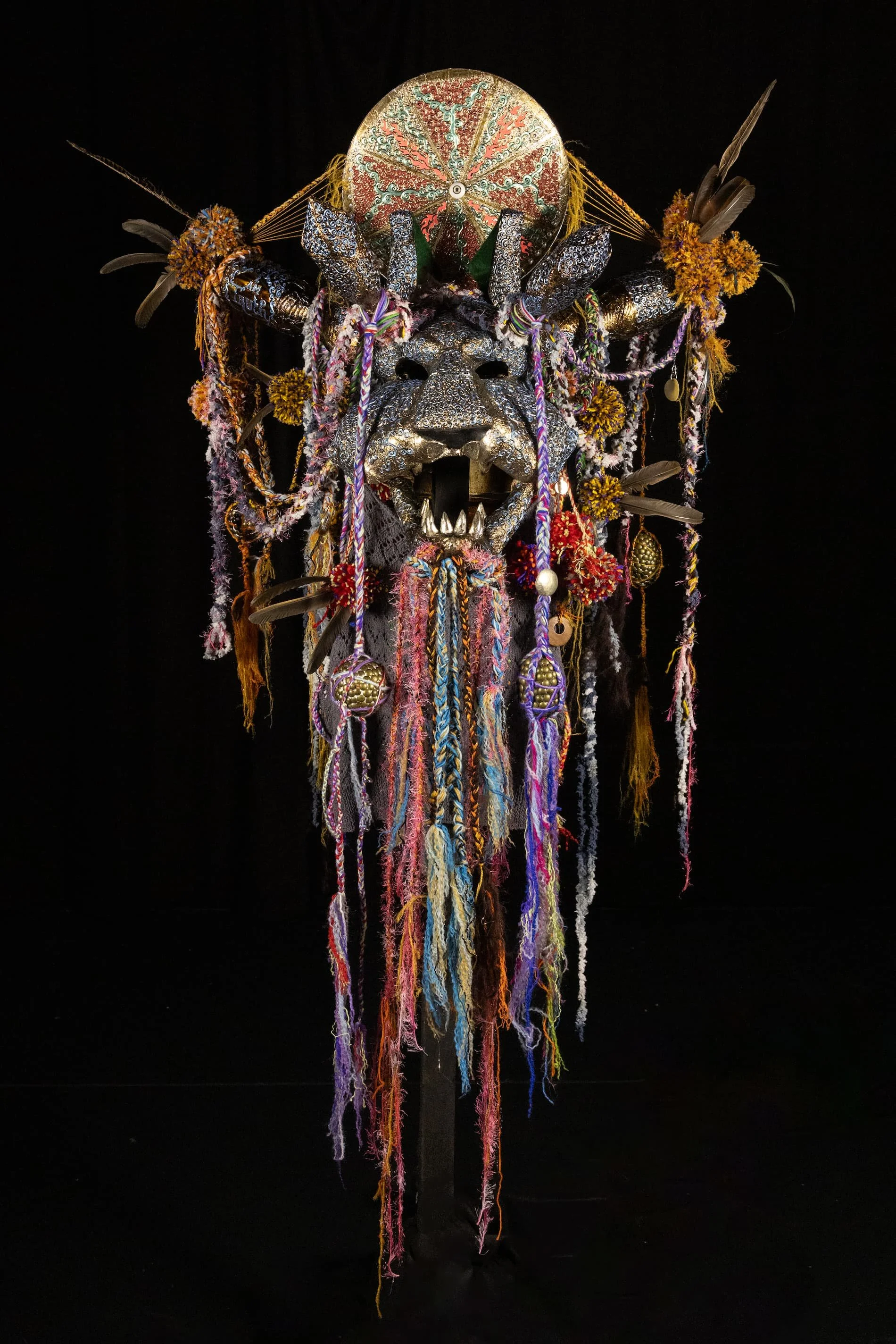 An elaborate tribal mask with a lion's face, decorated with feathers, beads, and colorful yarns, set against a black background.