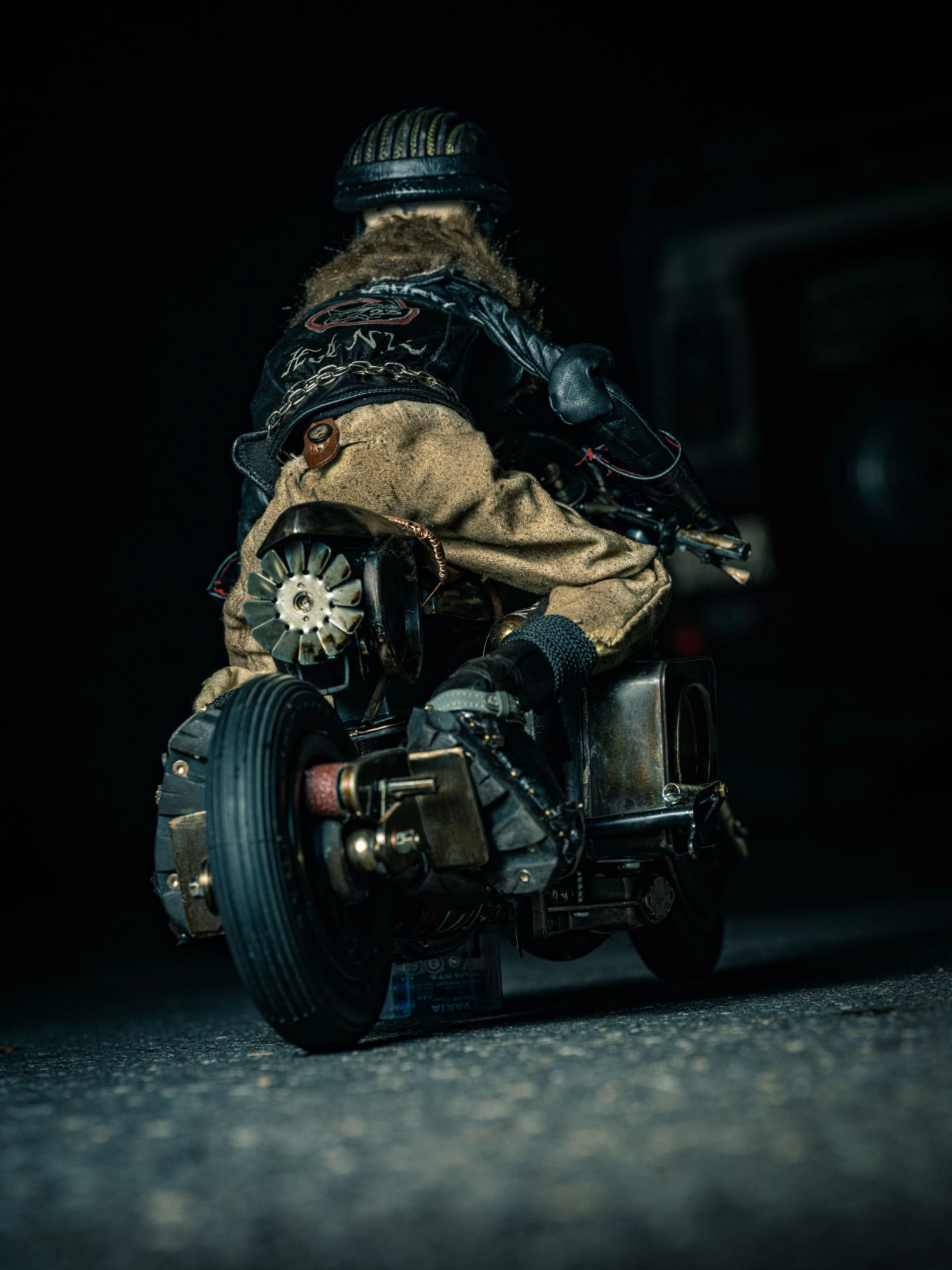 A miniature figure of a person riding a custom motorized wheelchair, wearing a beanie hat, leather jacket, and jeans, on a dark street at night.