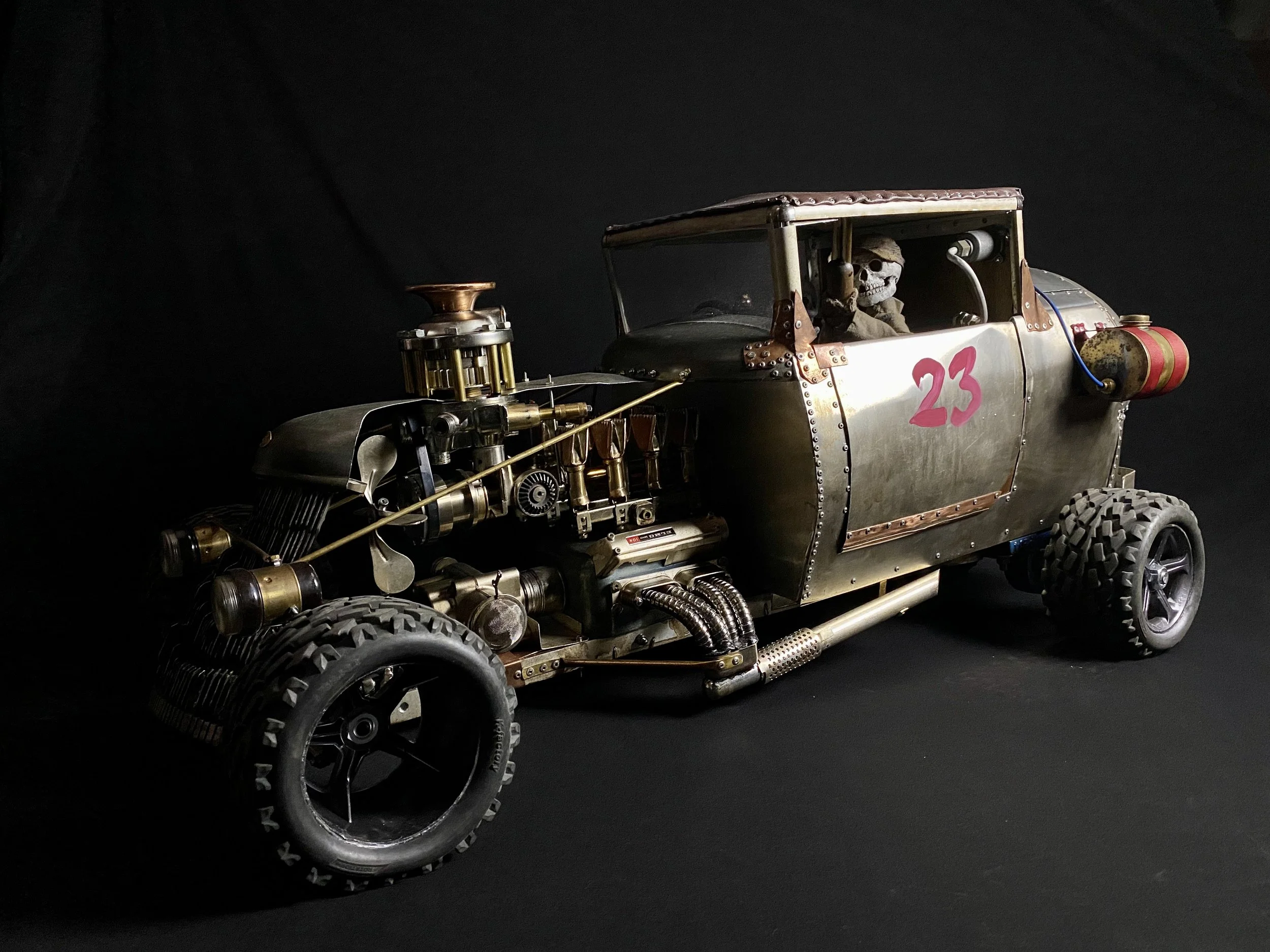 A detailed steampunk style miniature car with metallic body, exposed mechanical parts including pipes and gears, and the number 23 painted on the side. The interior is visible with a skeleton figure seated inside, against a dark background.
