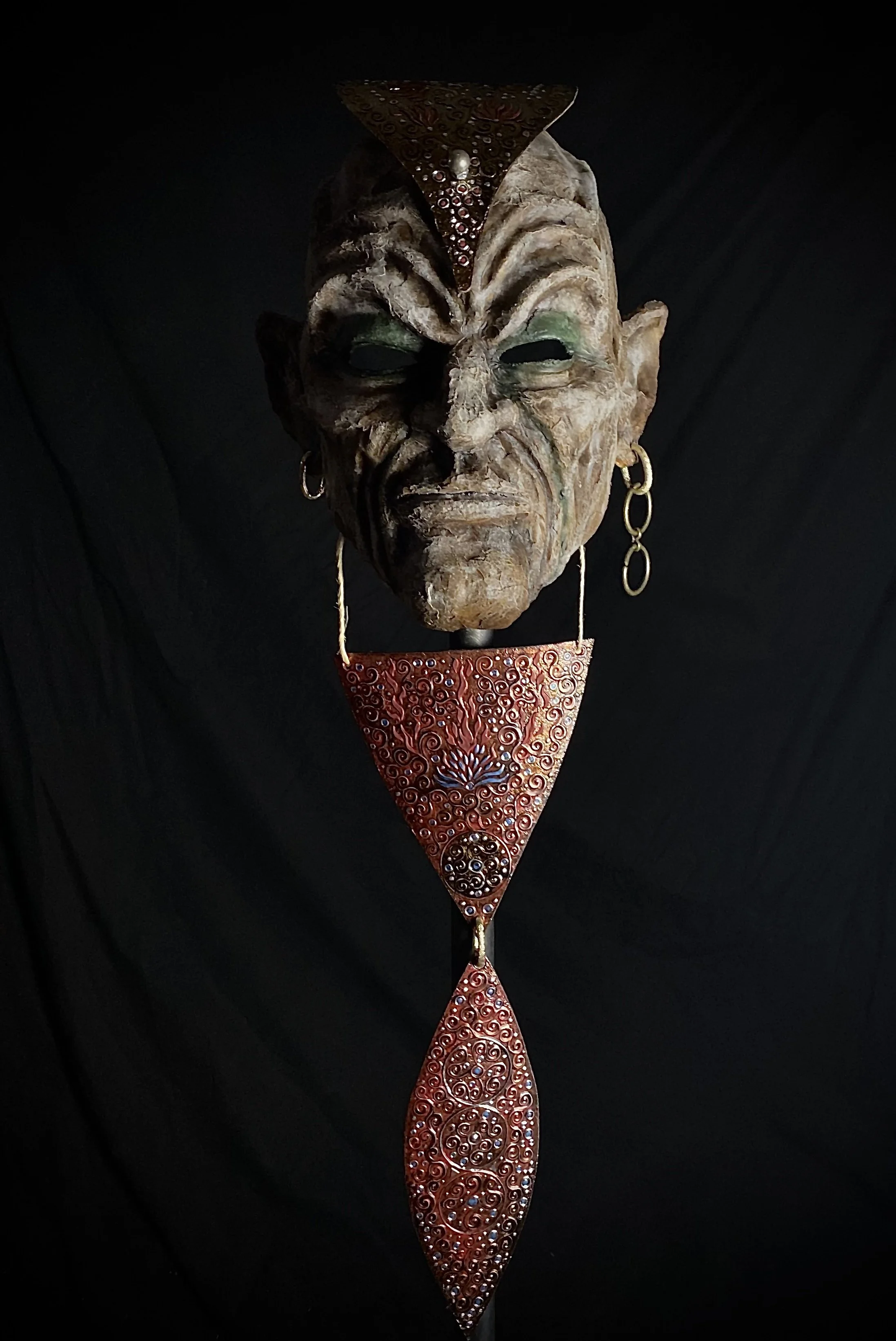 A tribal or artistic mask with ancient, weathered features, dark eye sockets, earrings, and elaborate red and gold jewelry hanging below.