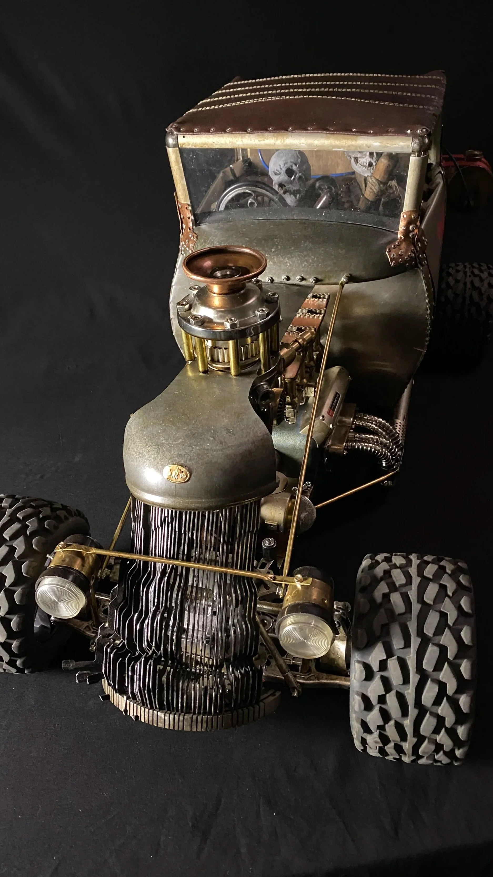 A miniature model of a vintage car with a metal body, large textured tires, and skull decorations inside the windshield.