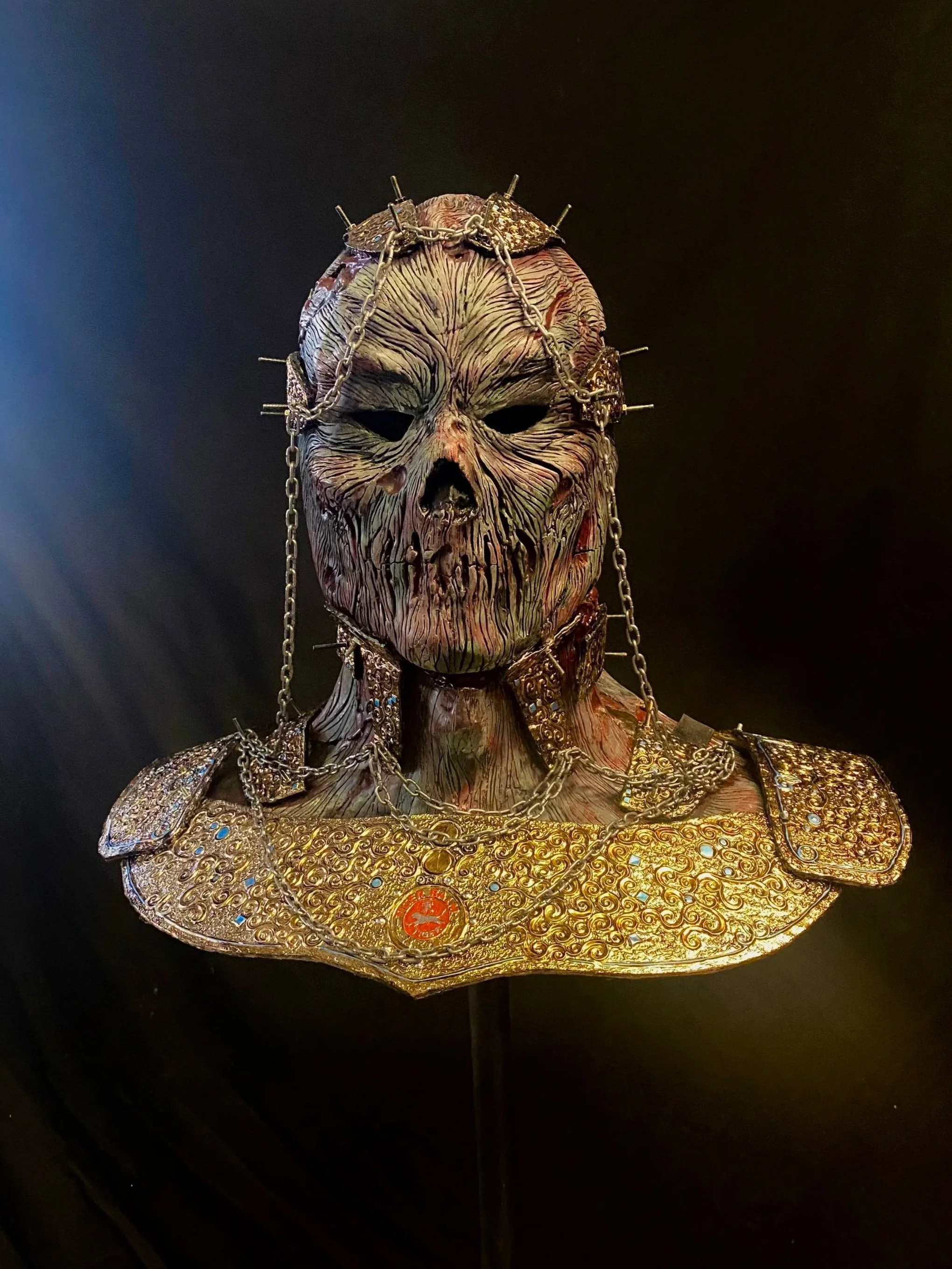 A detailed mask with a skull-like face, textured skin, and hollow eyes, adorned with chains and gold jewelry, displayed against a dark background.