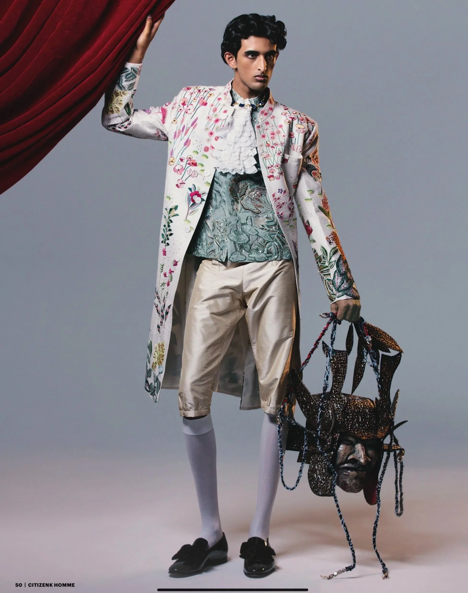 Man wearing an intricately embroidered long coat, ornate shirt, beige knee-length shorts, white socks, and black shoes, holding a decorative mask with chains and colorful embellishments, standing against a plain backdrop.