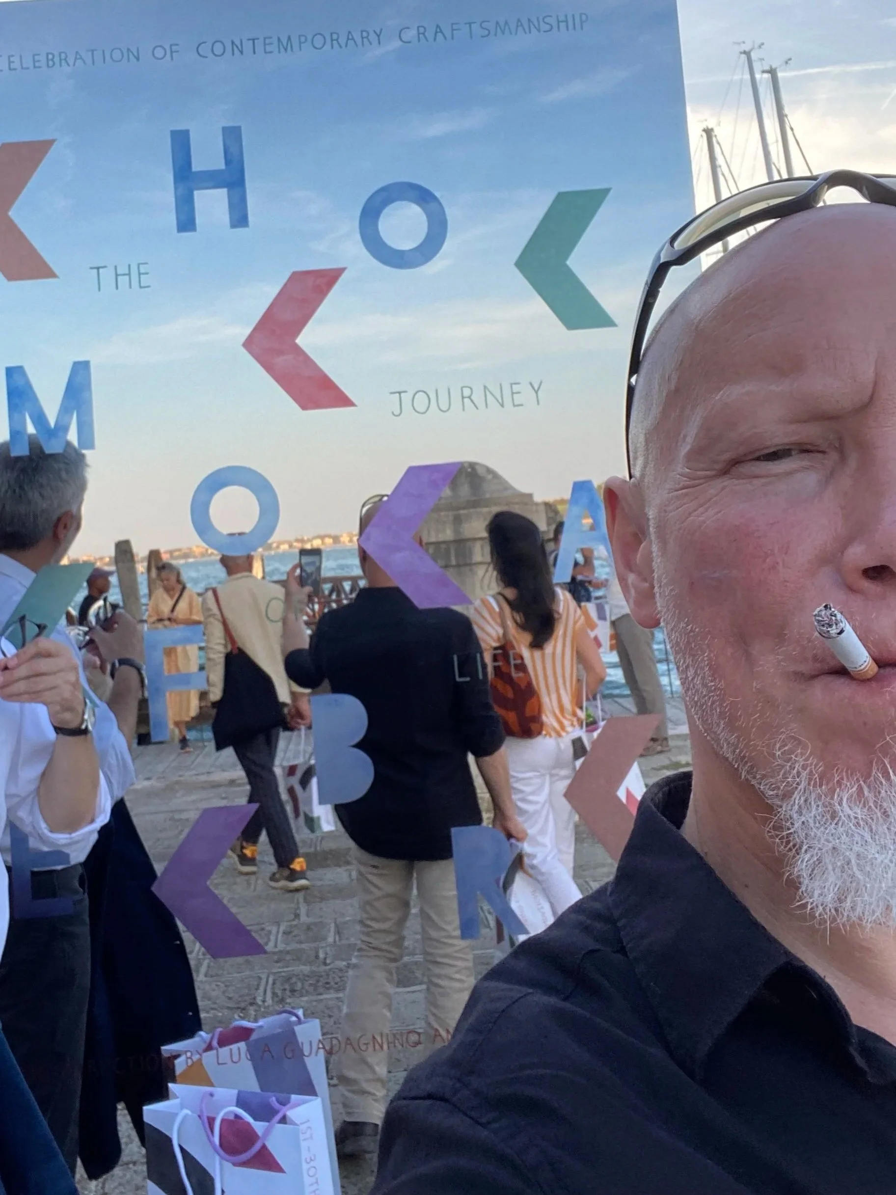 A person takes a selfie at an outdoor event near a body of water. The background is a colorful sign with the words 'Celebration of Contemporary Craftsmanship,' 'The Homer Journey,' and 'Life,' with people standing and taking photos.