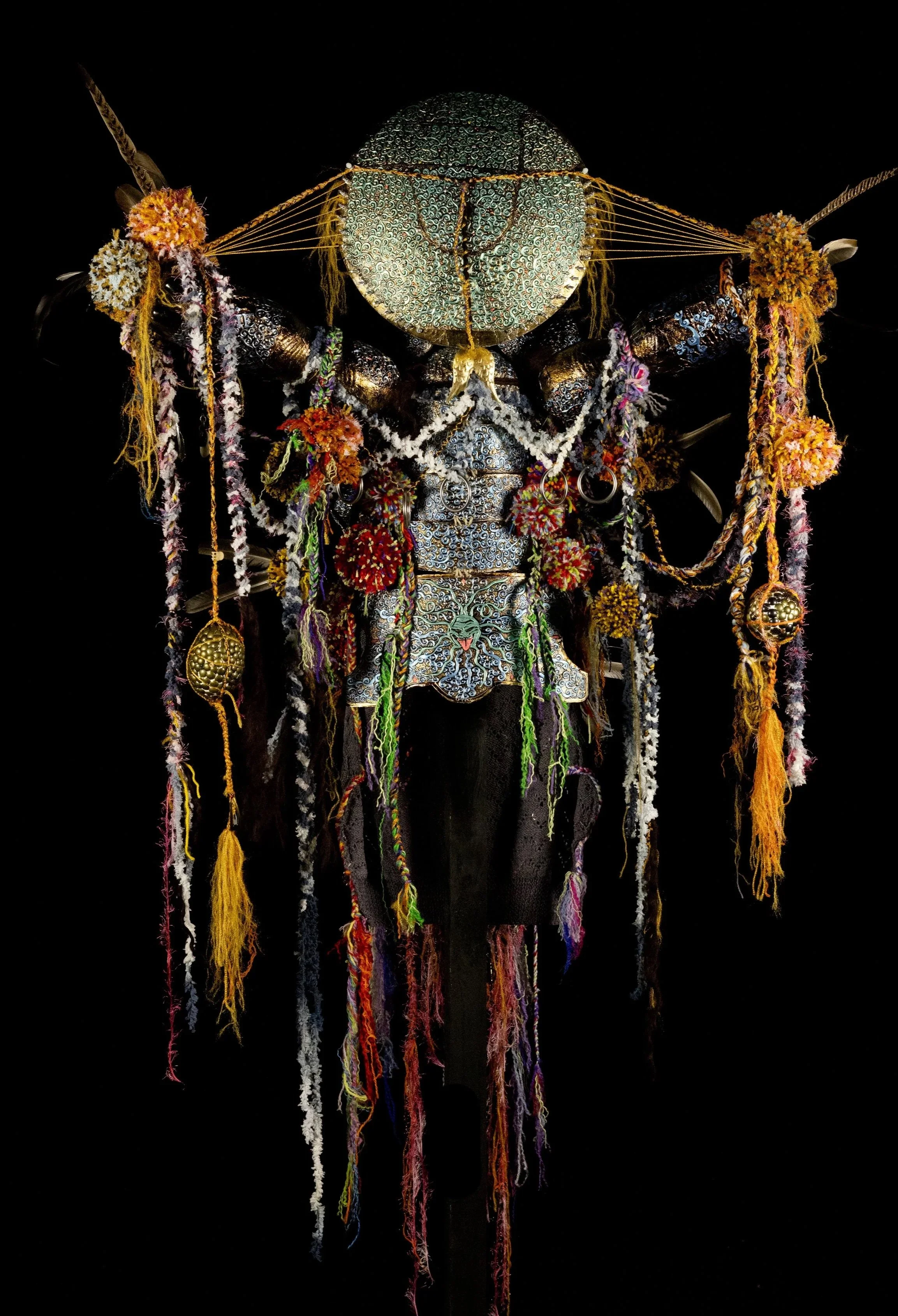 An intricate, colorful, textile and metal art piece resembling an abstract humanoid figure against a black background.