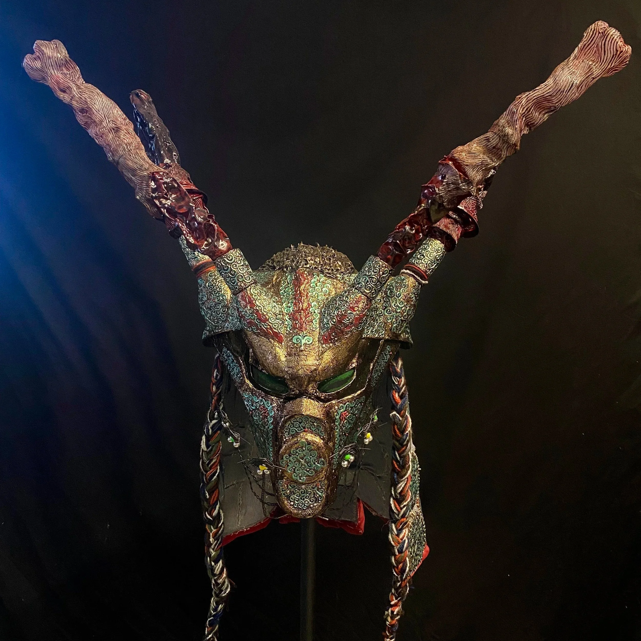 Decorative mask resembling a mythical creature with green eyes, large twisted horns, intricate patterns, and braided adornments hanging from the sides.