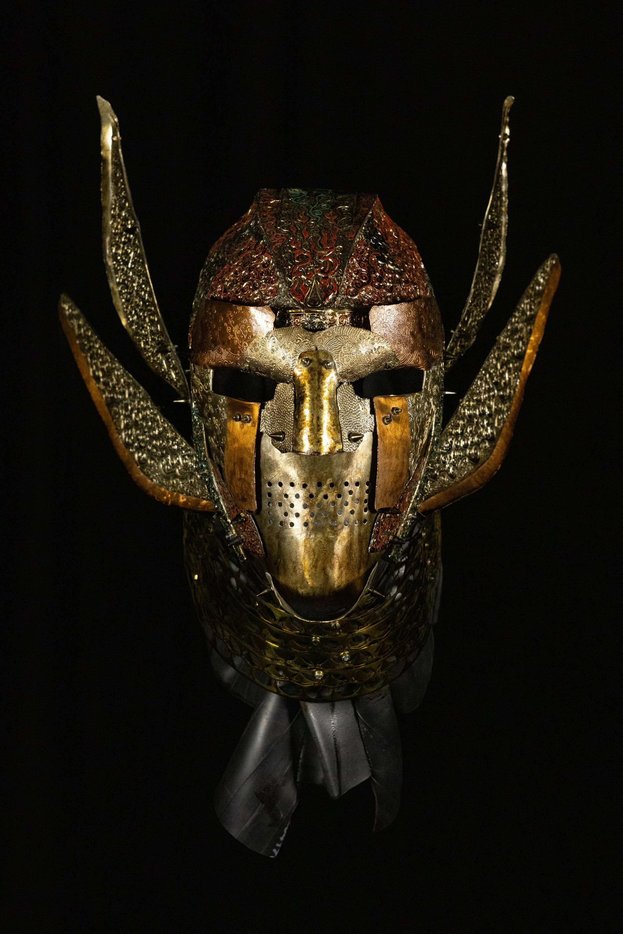 A metal sculpture of a mask, stylized insect or alien face, with intricate detailing and metallic textures, set against a black background.