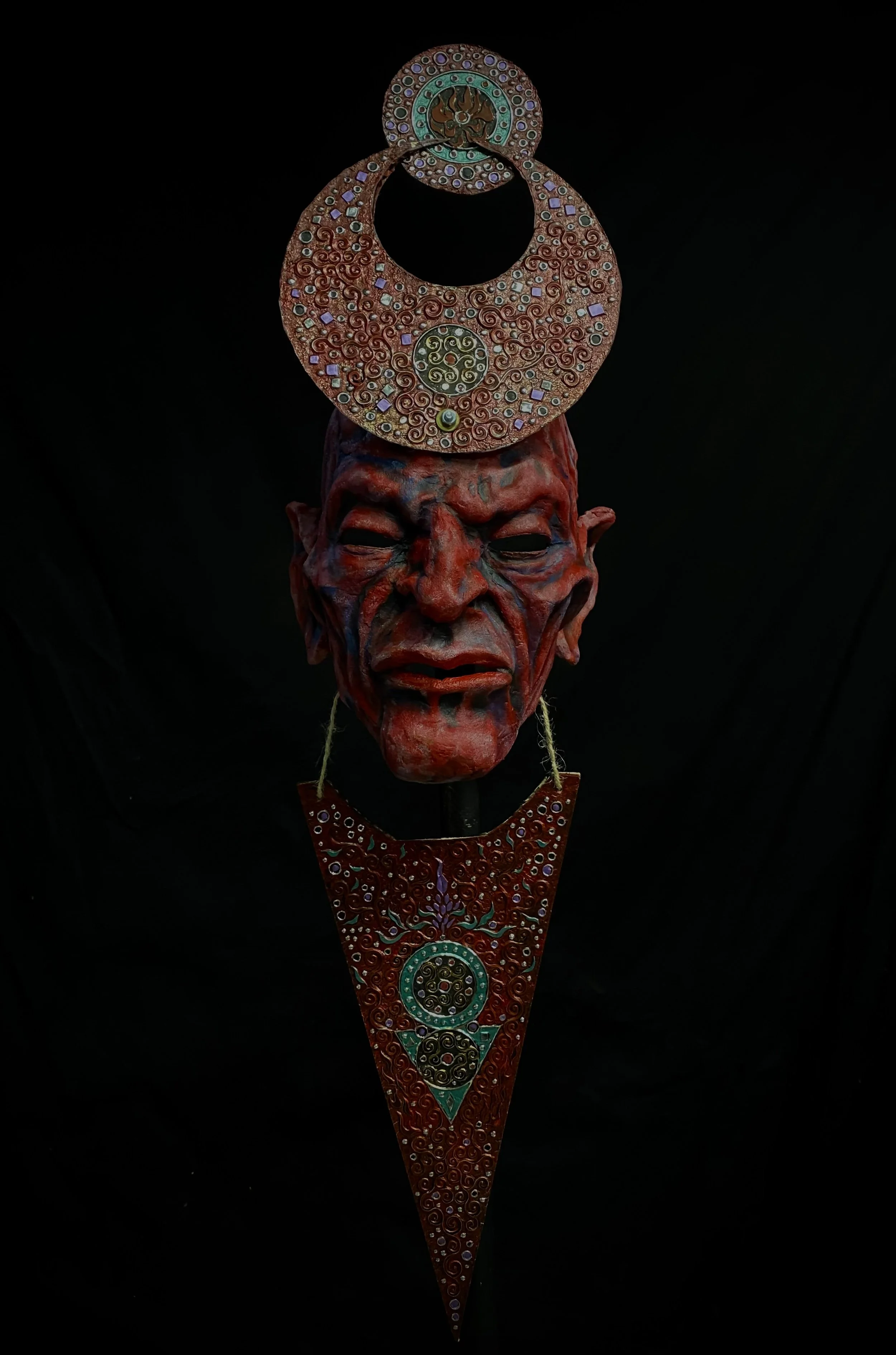 A display featuring a dark background with a red, tribal-looking mask, and attached decorative circular and triangular ornaments with intricate patterns and colorful designs.
