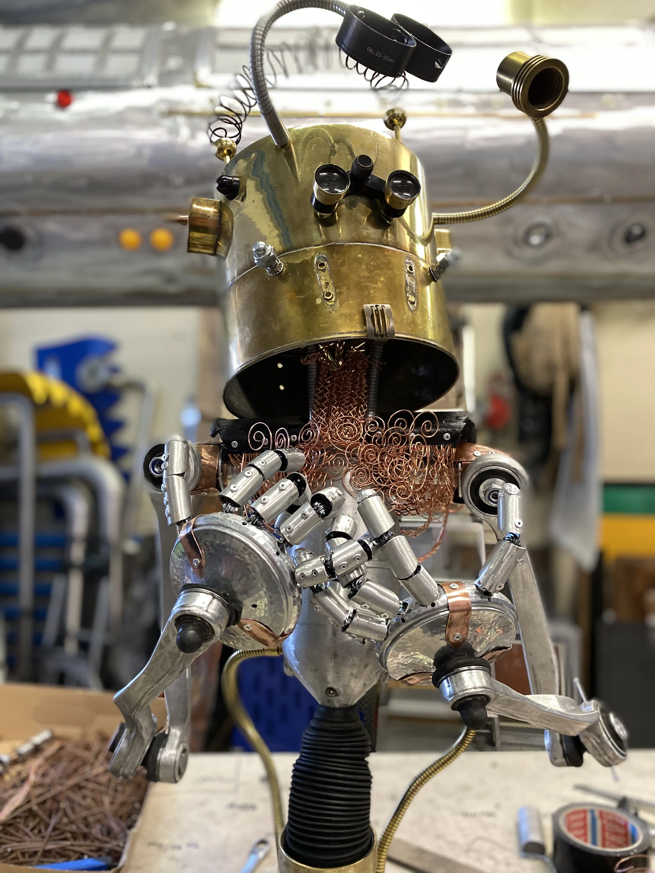 Close-up of a robot under construction with a brass head, metal limbs, and copper wiring, in a workshop.
