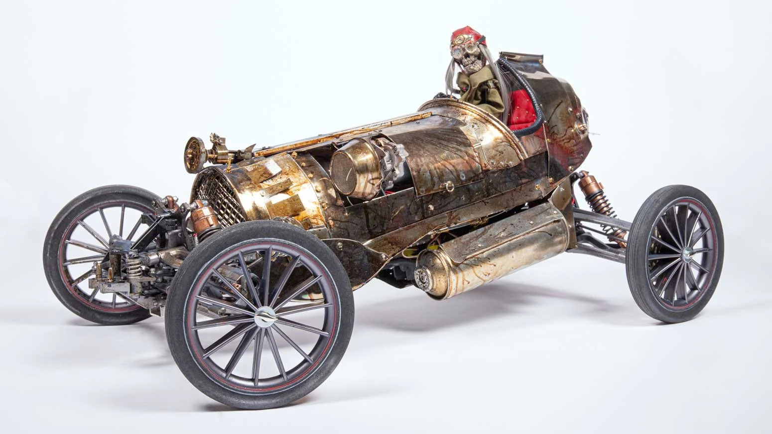 A vintage race car made of metal with an exposed engine, large spoked wheels, and a small cockpit with a skeleton figure wearing a helmet sitting inside.