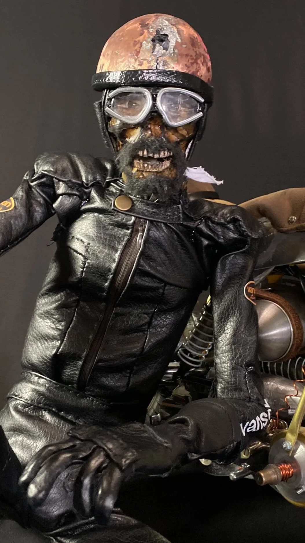 A Halloween skeleton dressed as a biker, wearing a black leather jacket, gloves, and vintage goggles, with a space-themed moon photo as the helmet, sitting on a motorcycle.