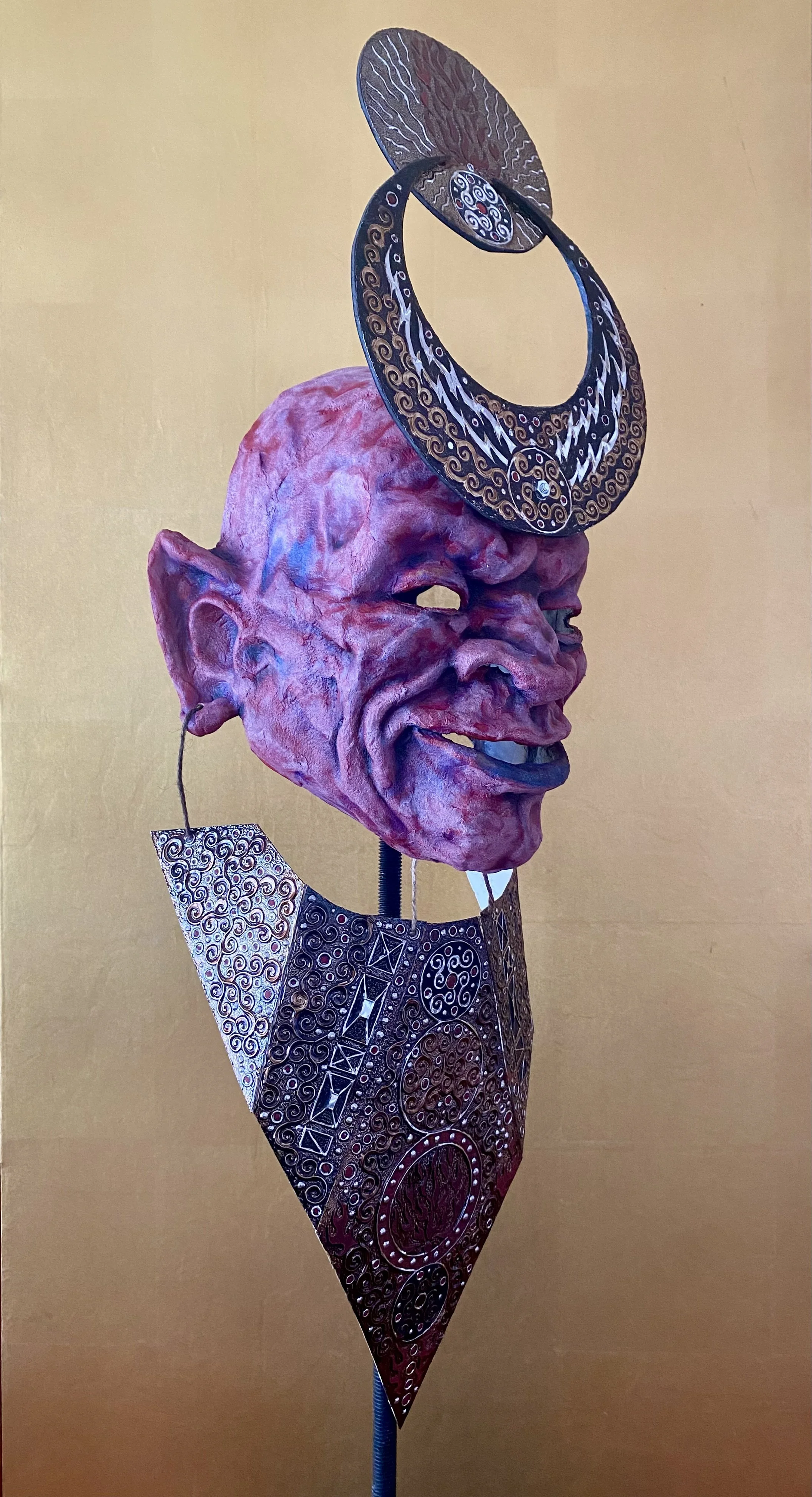 A sculpture featuring a large, exaggerated, humanoid face with wrinkled, pinkish skin and a big nose, wearing a metallic, decorated earring. The face has a wide, toothy grin and is topped with an ornate, circular metallic headpiece and a decorative n