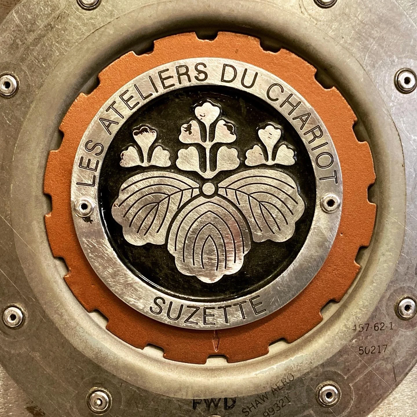 Close-up of a metal emblem with a butterfly design, inscribed with the words 'Les Ateliers du Charriol,' 'Suzette,' and some technical markings and screws around the edges.