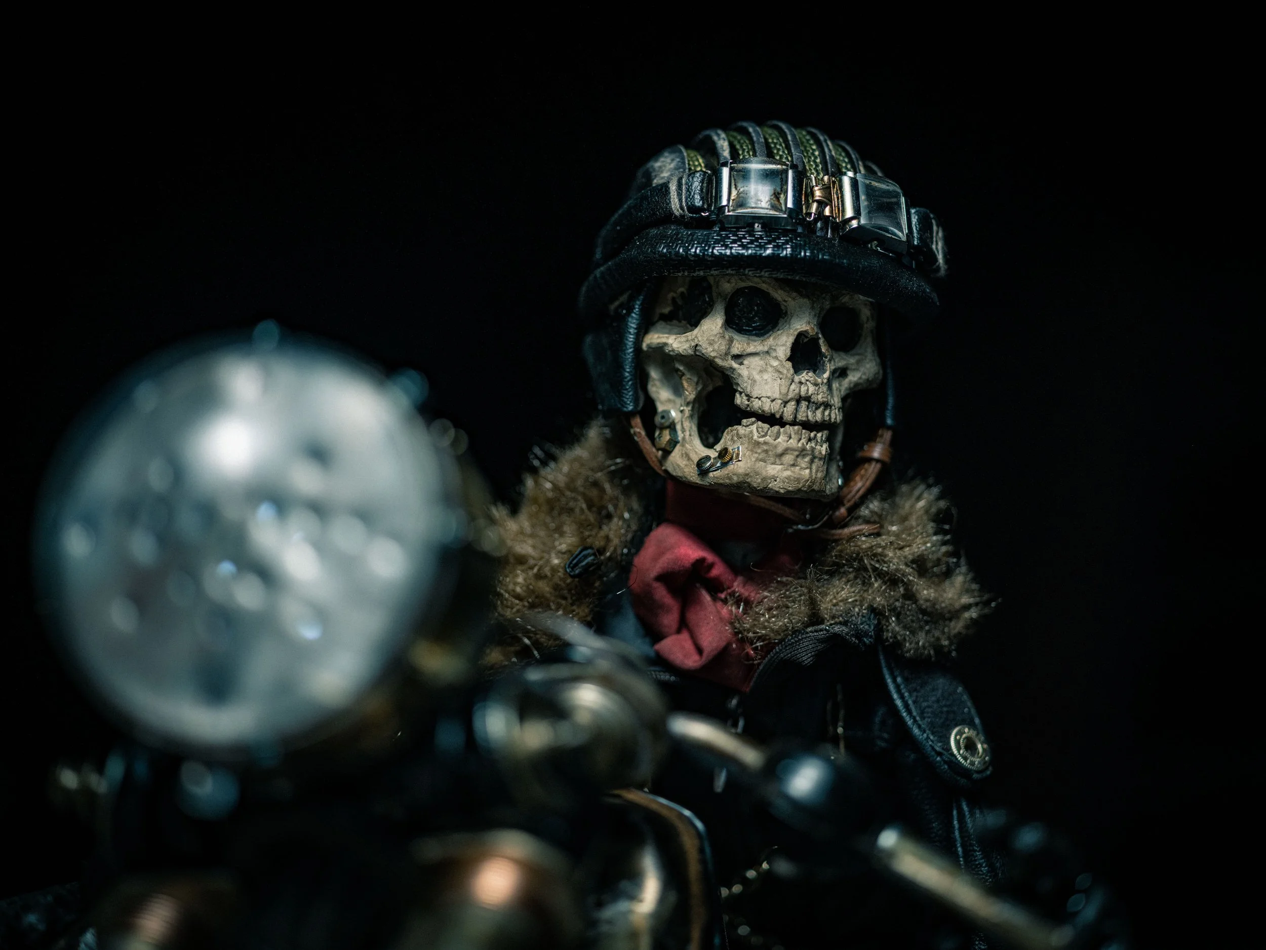 A skeleton wearing a motorcycle helmet and jacket, sitting on a motorcycle with the headlight in the foreground, against a black background.