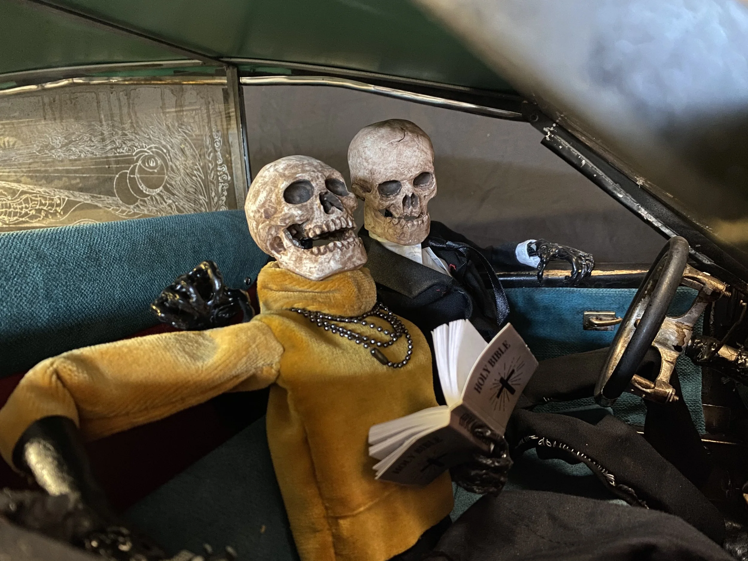 Skeleton figures dressed as a driver and passenger, sitting in a vintage car, with the passenger reading a Bible and wearing a yellow jacket and necklaces.
