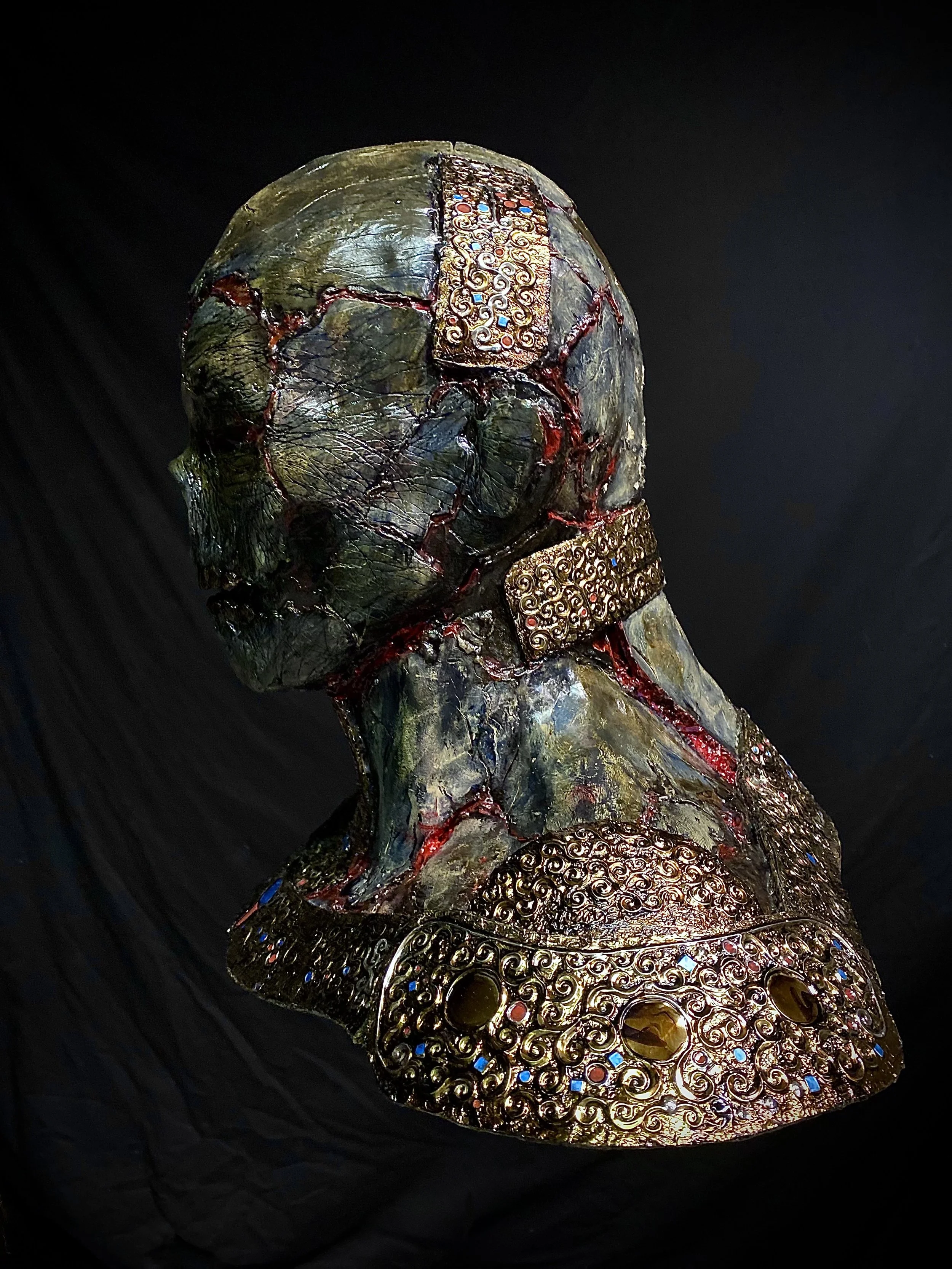 A sculptural bust of a person with a textured, multicolored surface resembling cracked stone, decorated with intricate gold metalwork and gemstones. The sculpture is set against a black background.