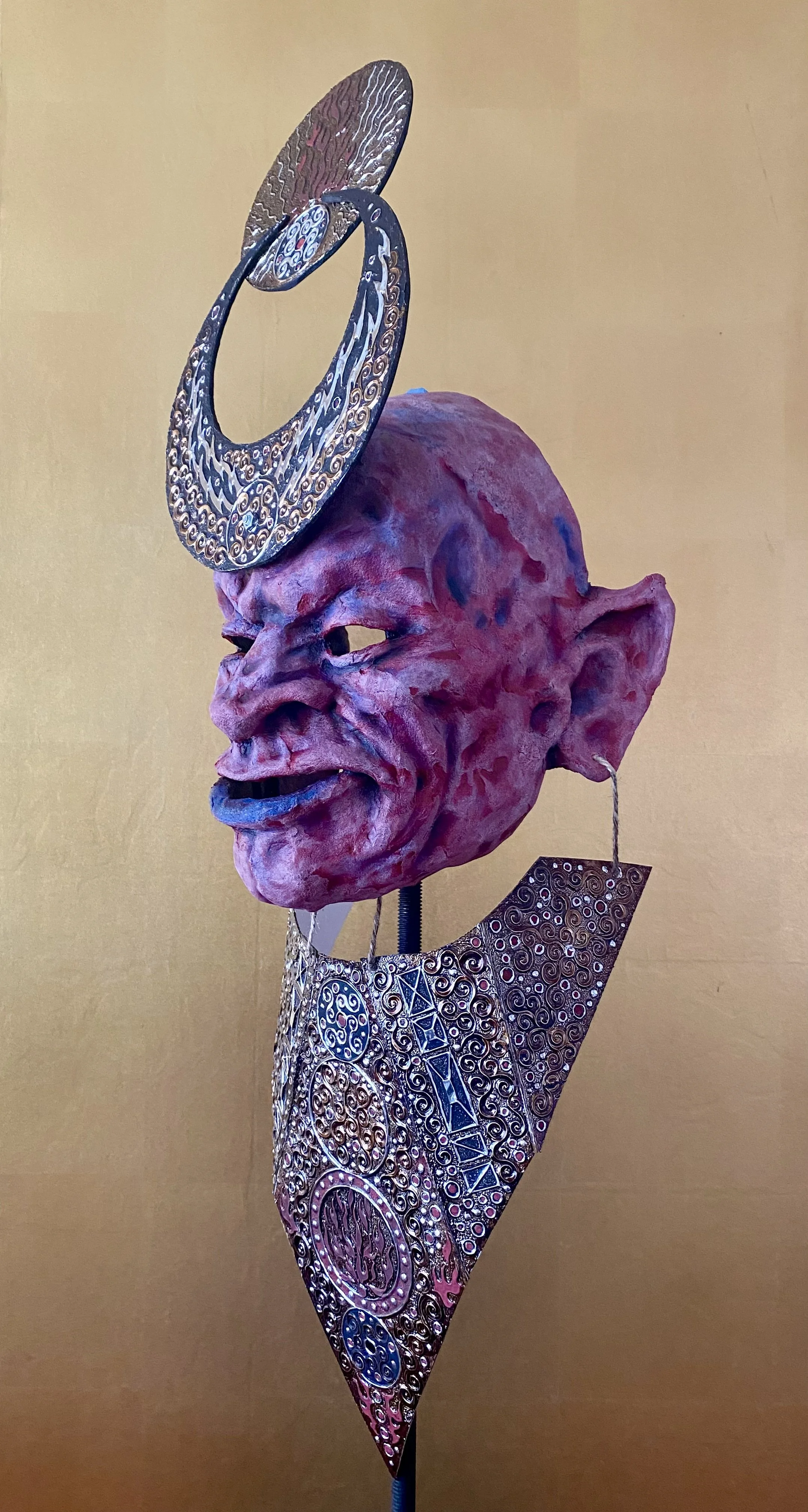 A sculpture of an artistic, exaggerated red face with large ears, a prominent nose, and a wide mouth, adorned with decorative metallic jewelry and an ornate headpiece, set against a plain yellow background.