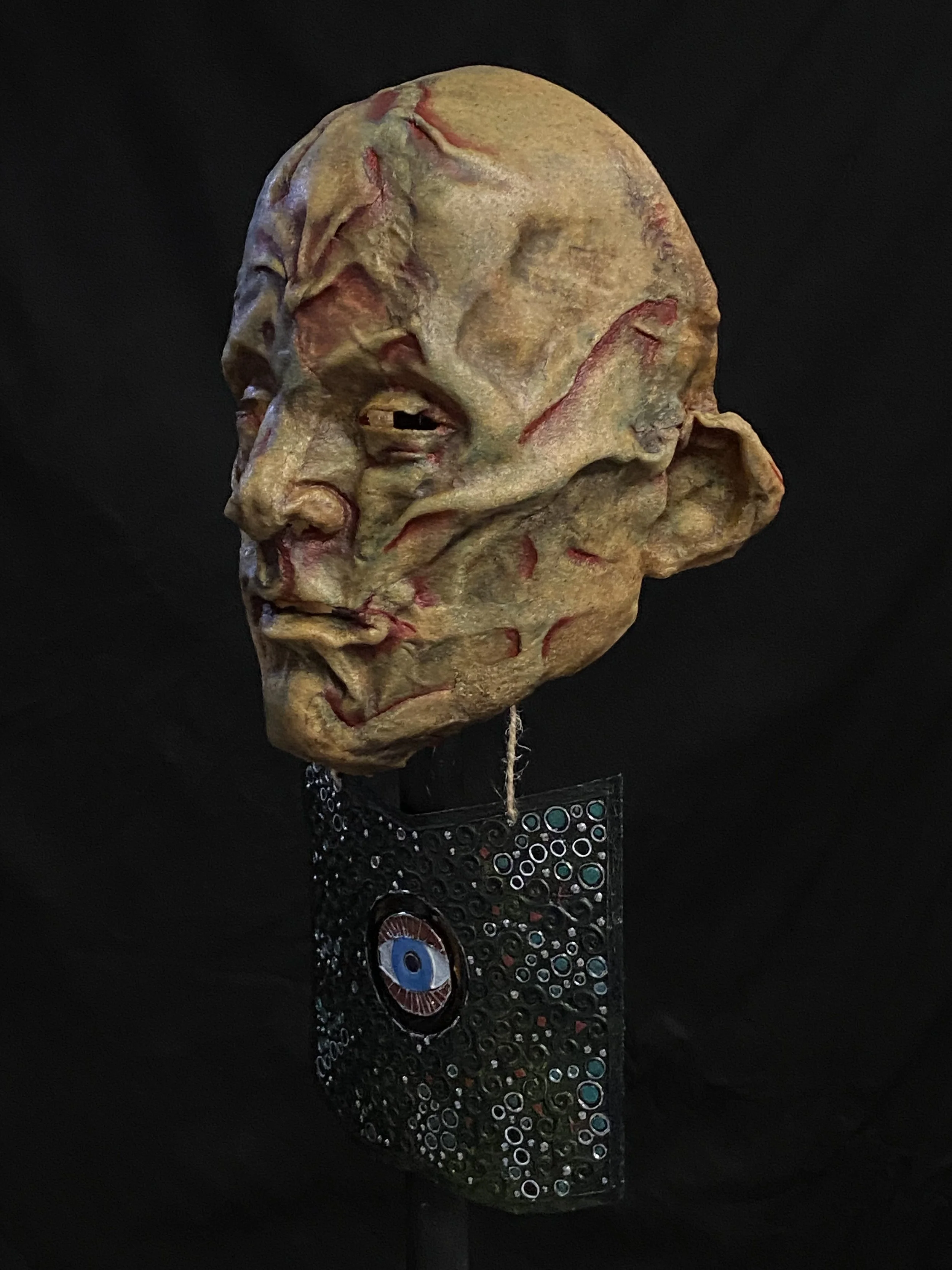 A surreal sculpture of a human face with a textured, worn appearance, mounted on a decorative black stand with circular patterns and an eye design.