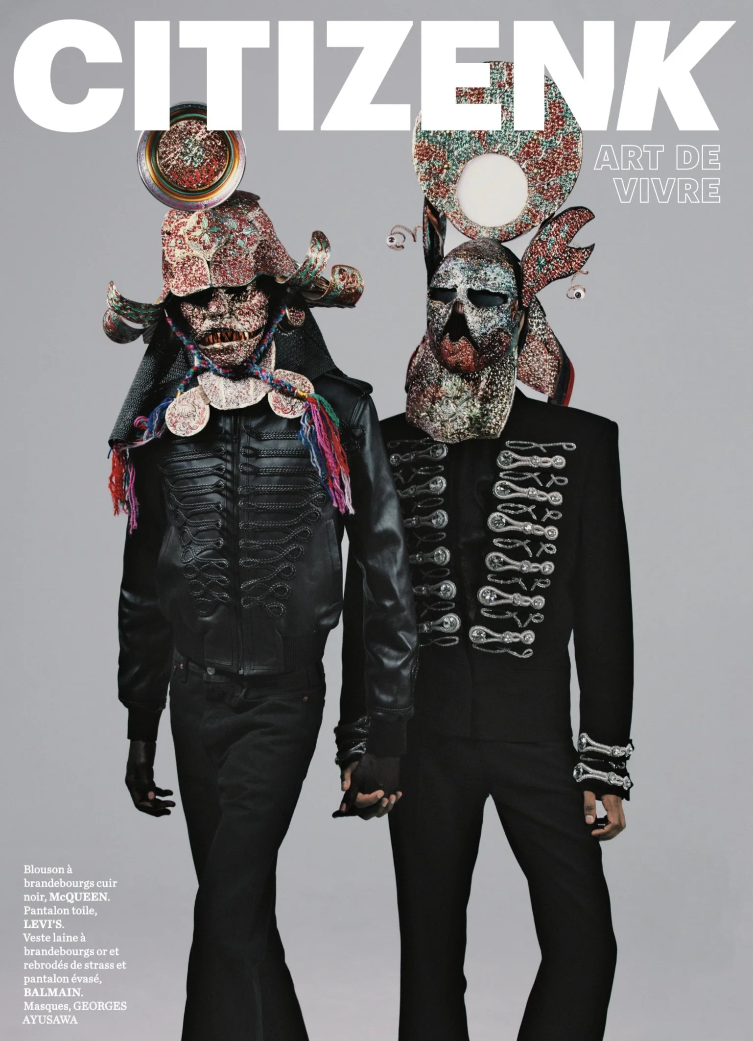Two individuals wearing black leather jackets with silver embroidery and mask-like headpieces featuring mixed textures, colorful patterns, and abstract designs, holding hands. The background is plain and light gray, a cover from CITIZEN K magazine