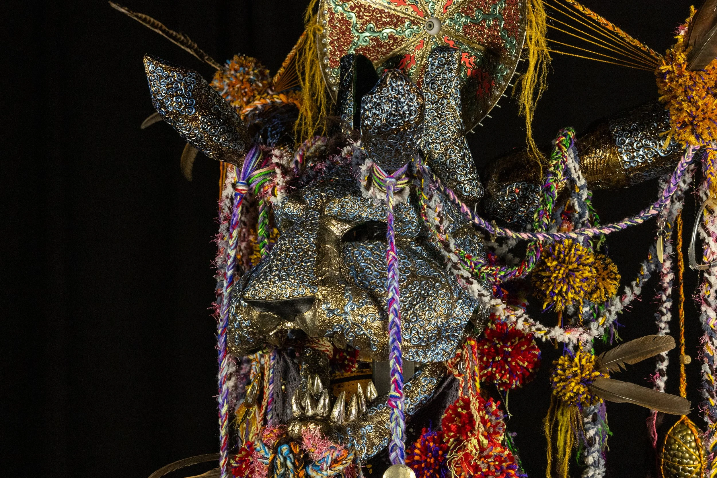 Close-up of a decorative mask resembling a lion or feline, adorned with bright, colorful textiles, beads, sequins, lace, feathers, and ropes, set against a black background.