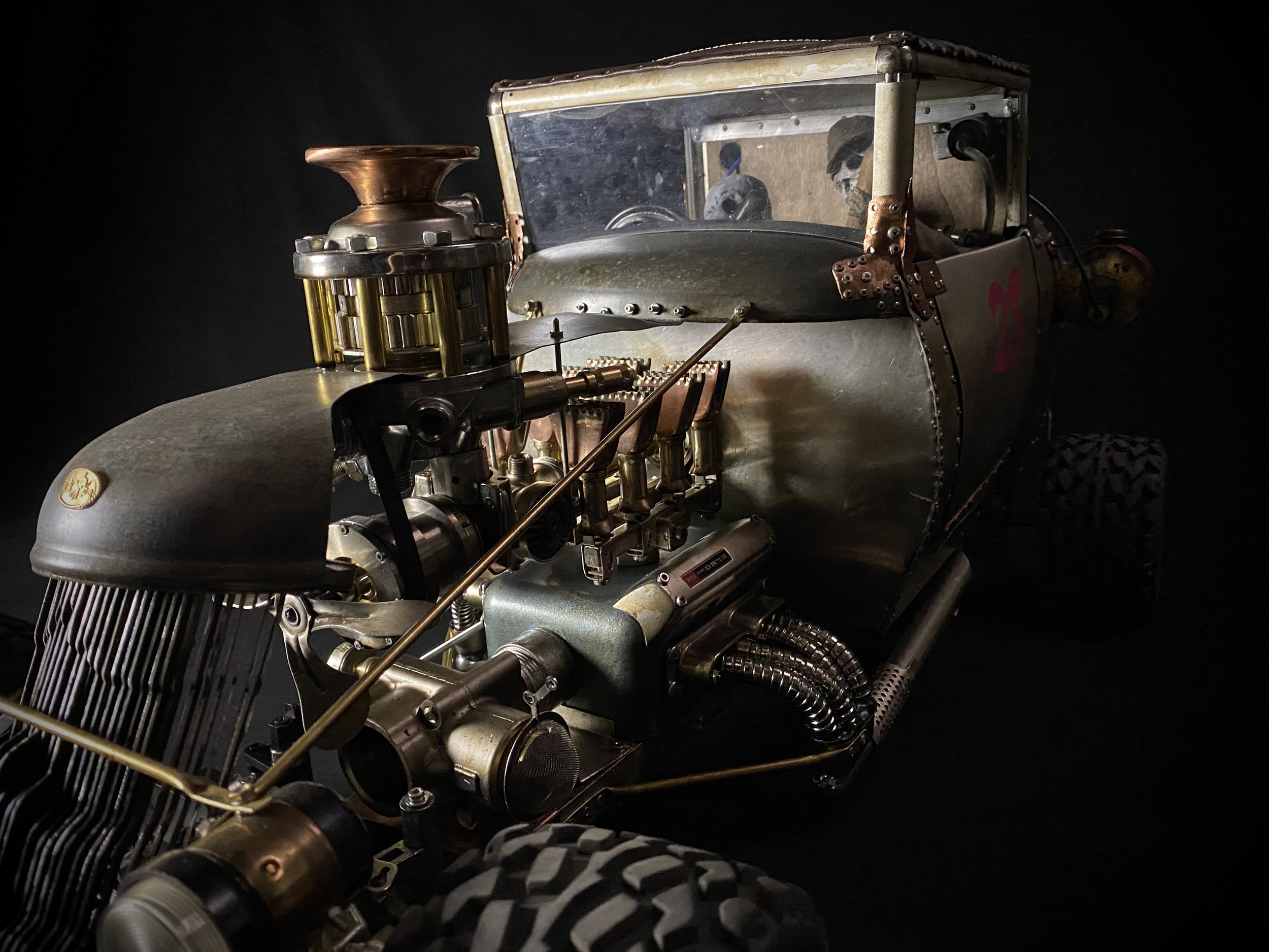 Close-up of a vintage hot rod with a metallic body, exposed engine components, and large tires, set against a dark background.