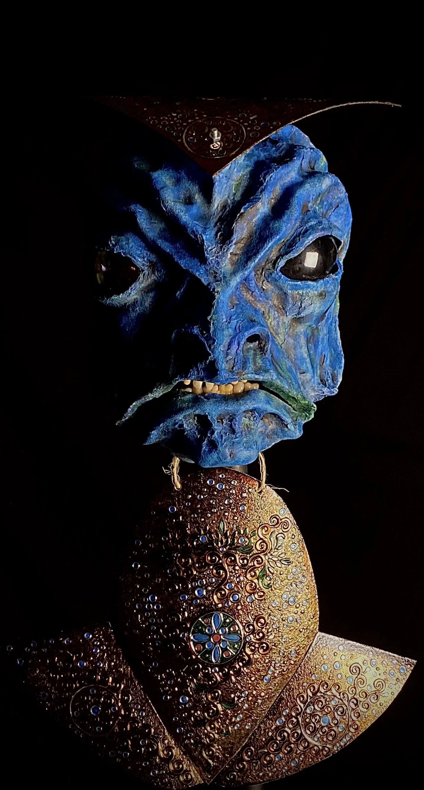 A decorative mask with a blue face, black eyes, and exaggerated facial features, complemented by an ornate, bejeweled pendant or necklace beneath it, set against a black background.