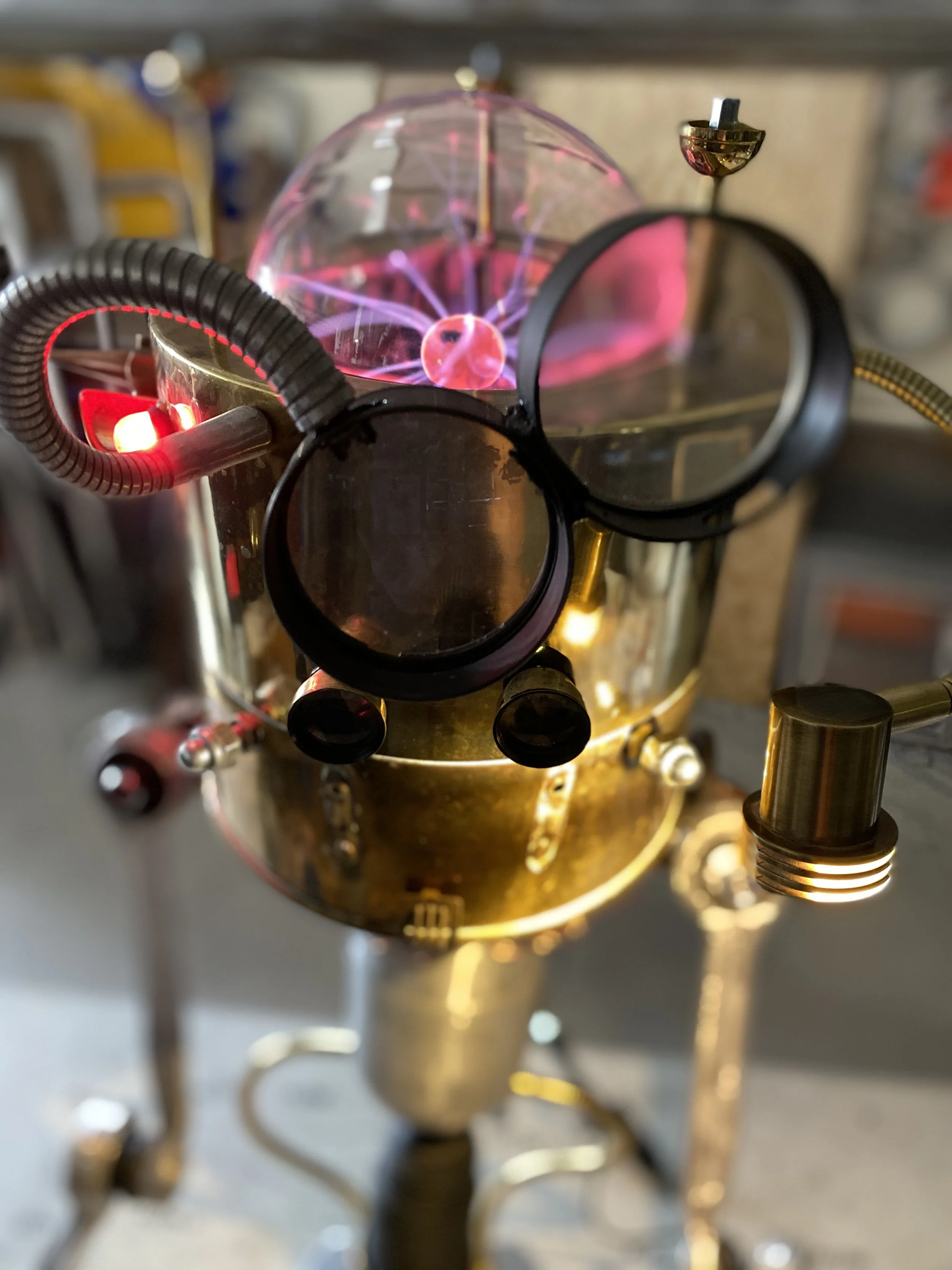 Close-up of a Tesla coil with electrical arcs and sparks, positioned behind a pair of safety goggles, in a workshop or laboratory setting.