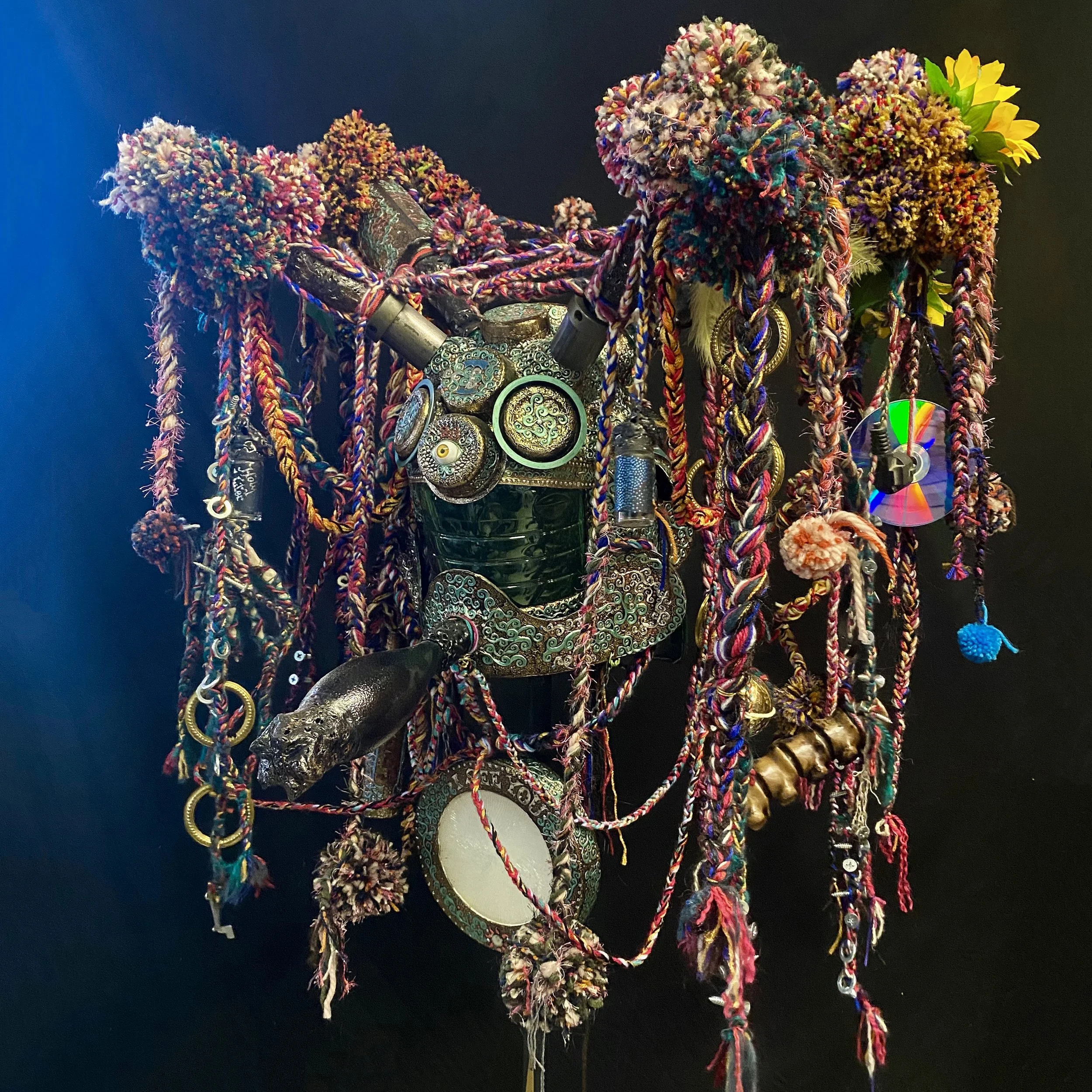 An eclectic art piece resembling a face, made from various colorful trinkets, jewelry, braided yarns, and found objects, set against a dark background.