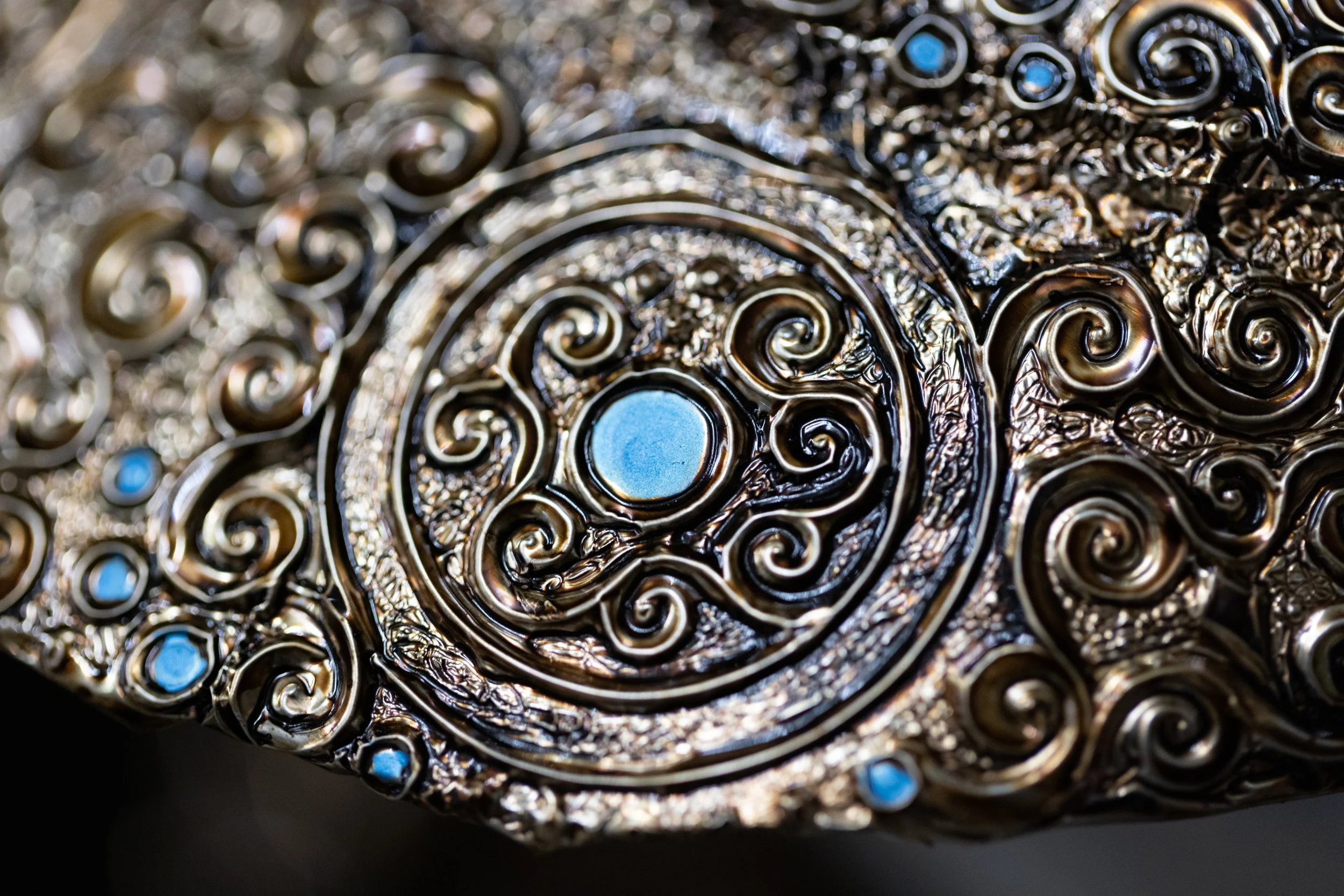 Close-up of an ornate, metallic object with intricate swirling patterns and small blue accents.