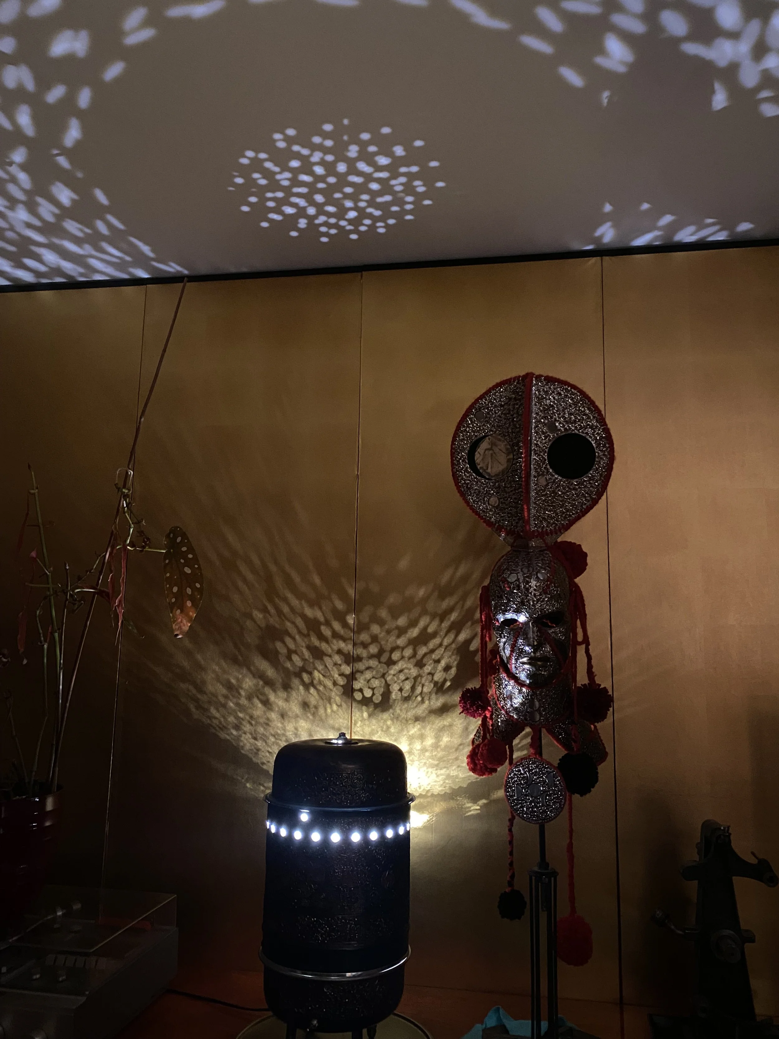 Decorative African mask with red accents, hanging on a wooden wall, illuminated by a lamp casting intricate shadows on the ceiling.