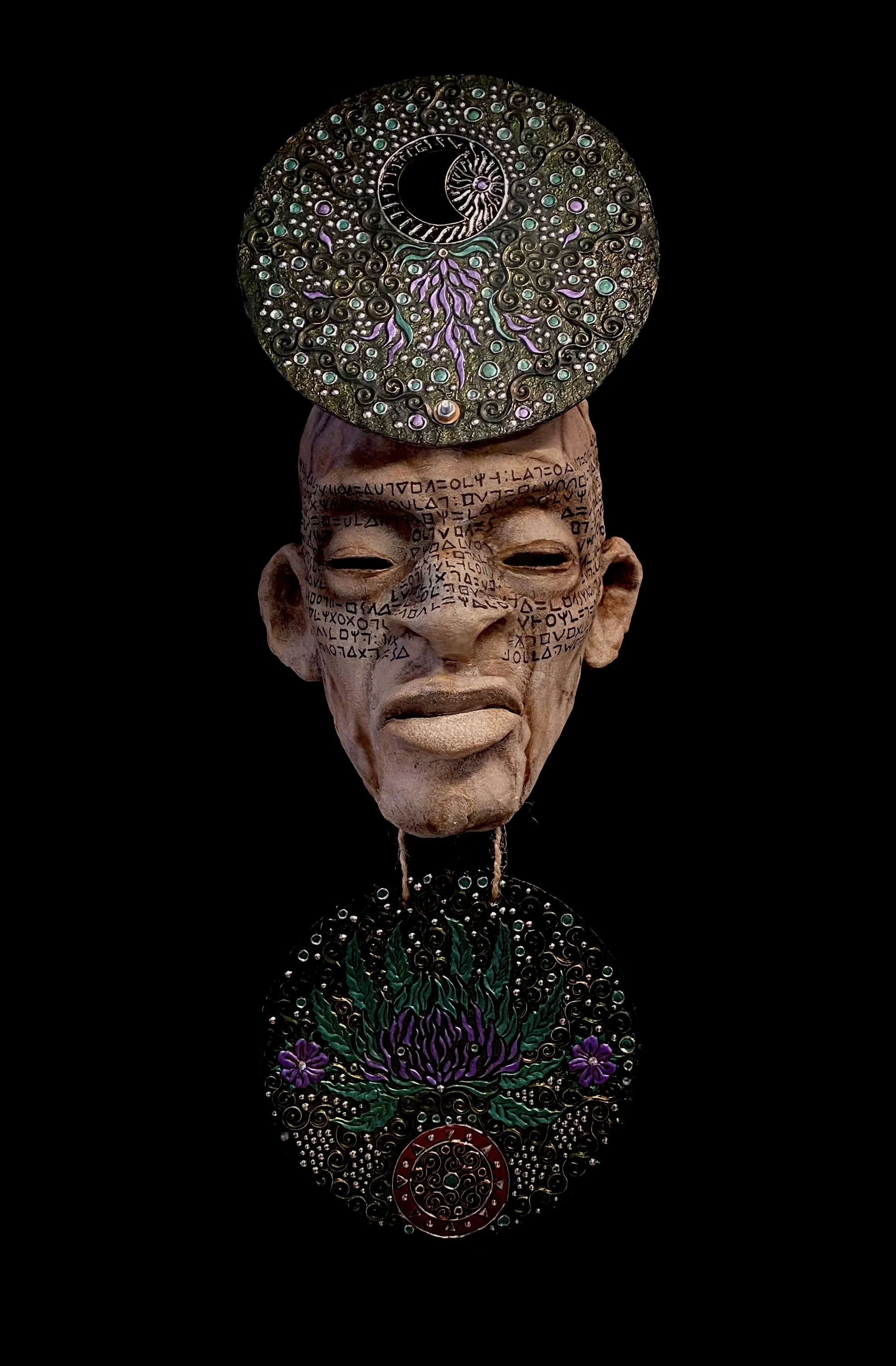 Mixed media sculpture featuring an elongated human face with inscriptions on the forehead, a neutral expression, surrounded by ornate disks with celestial and botanical motifs, against a black background.